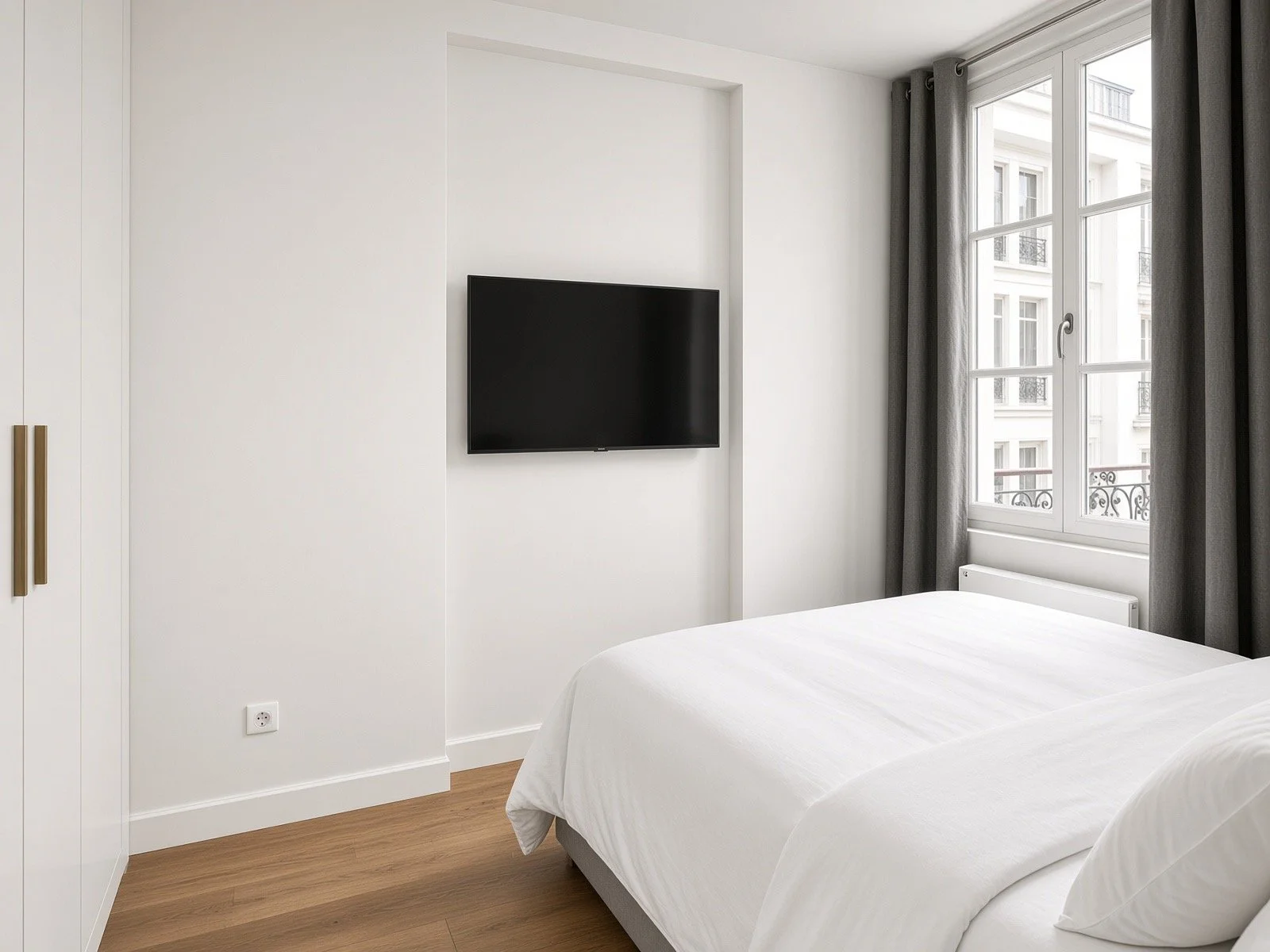 Minimalist bedroom with white walls, a mounted flat-screen TV, a window with gray curtains, a white bed with pillows, and wooden flooring.