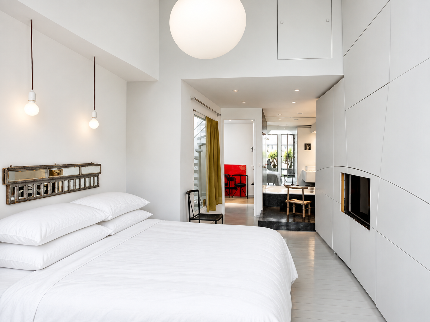 Modern bedroom with white bedding, minimalist decor, large circular ceiling light, and a split-level view into a living area with a black fireplace and plants.