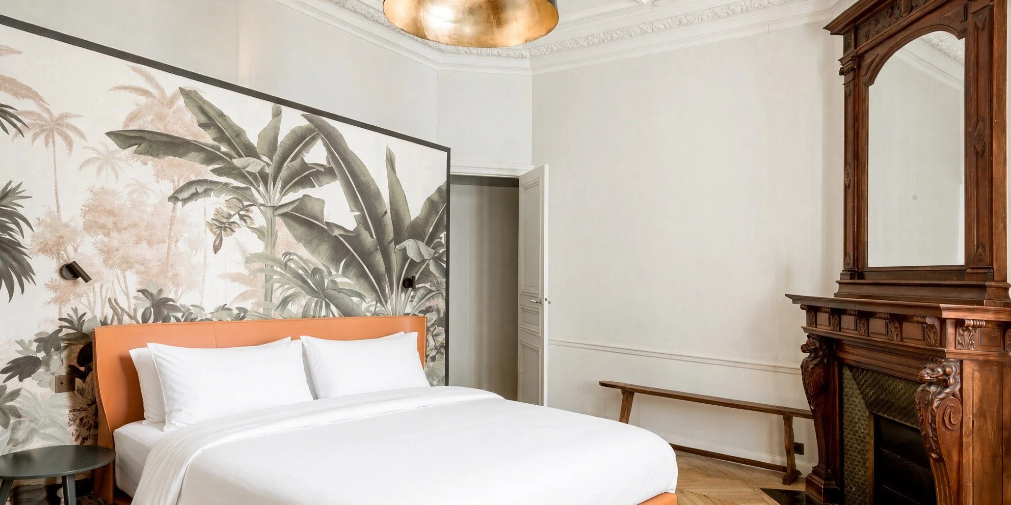 Bedroom with a bed, tropical wallpaper behind it, a small black side table, wooden mantel with mirror, wall-mounted reading lights, white walls and ceiling molding, and a partially open door.