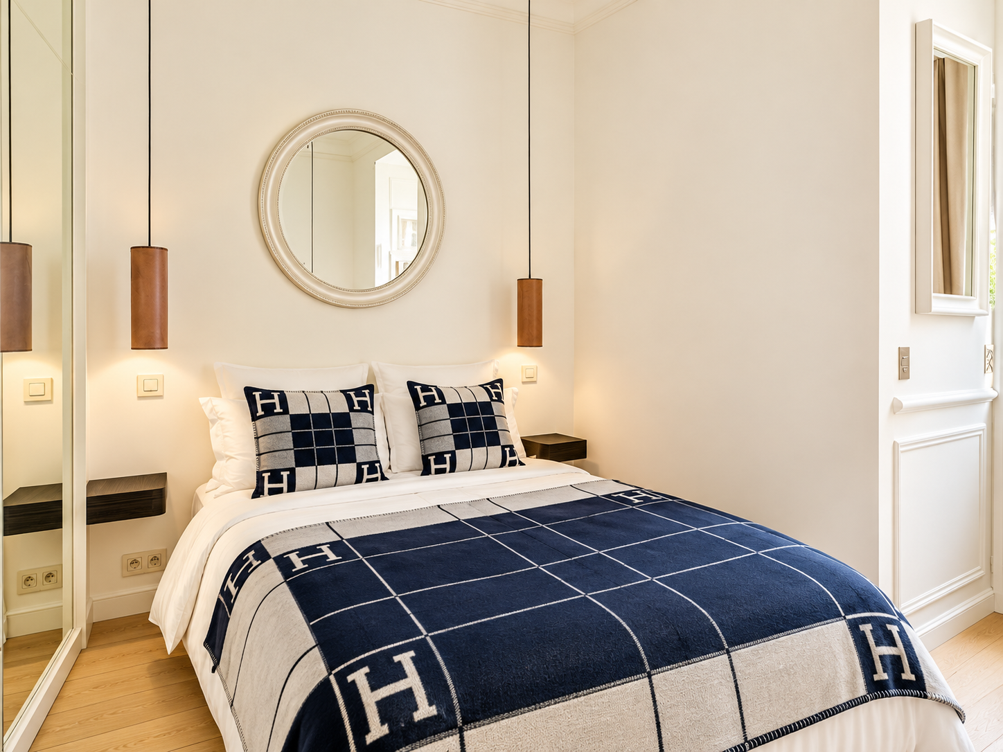 A neatly made bed with a navy blue and white plaid bedspread, featuring the letter 'H' on pillows and blanket, in a well-lit bedroom with a round mirror above the headboard, beige walls, and hanging pendant lights on either side.