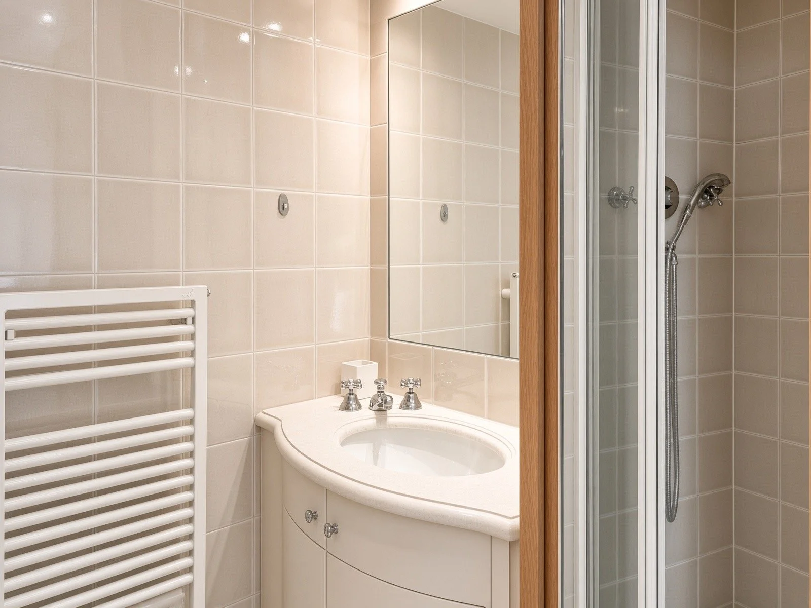 A small bathroom with beige tiled walls, a white mirror, a bathtub with a rounded corner, a white radiator, and a separate shower stall with a wooden door.