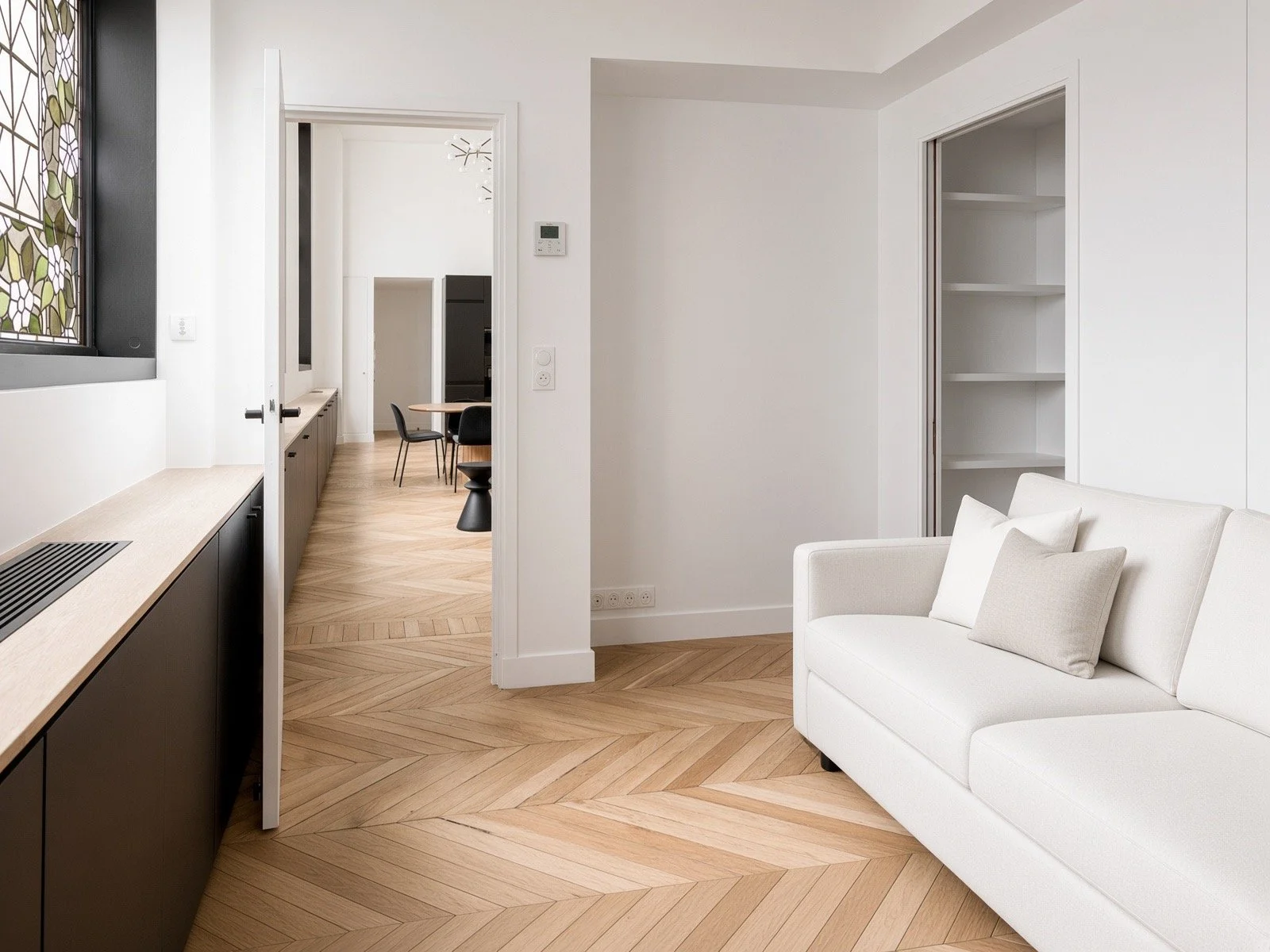 Modern living room with white walls, a cream sofa with pillows, and wood herringbone flooring, connected to a dining area with black chairs.