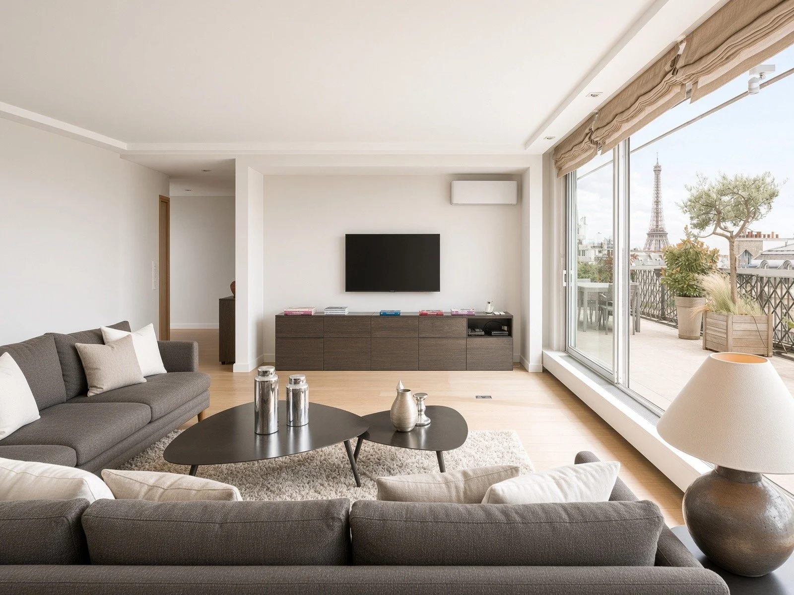 Modern living room with a gray sectional sofa, white and beige pillows, a black coffee table with decorative silver items, a wall-mounted TV, a wooden console, large glass sliding doors leading to a balcony with plants, and a view of the Eiffel Tower in Paris.