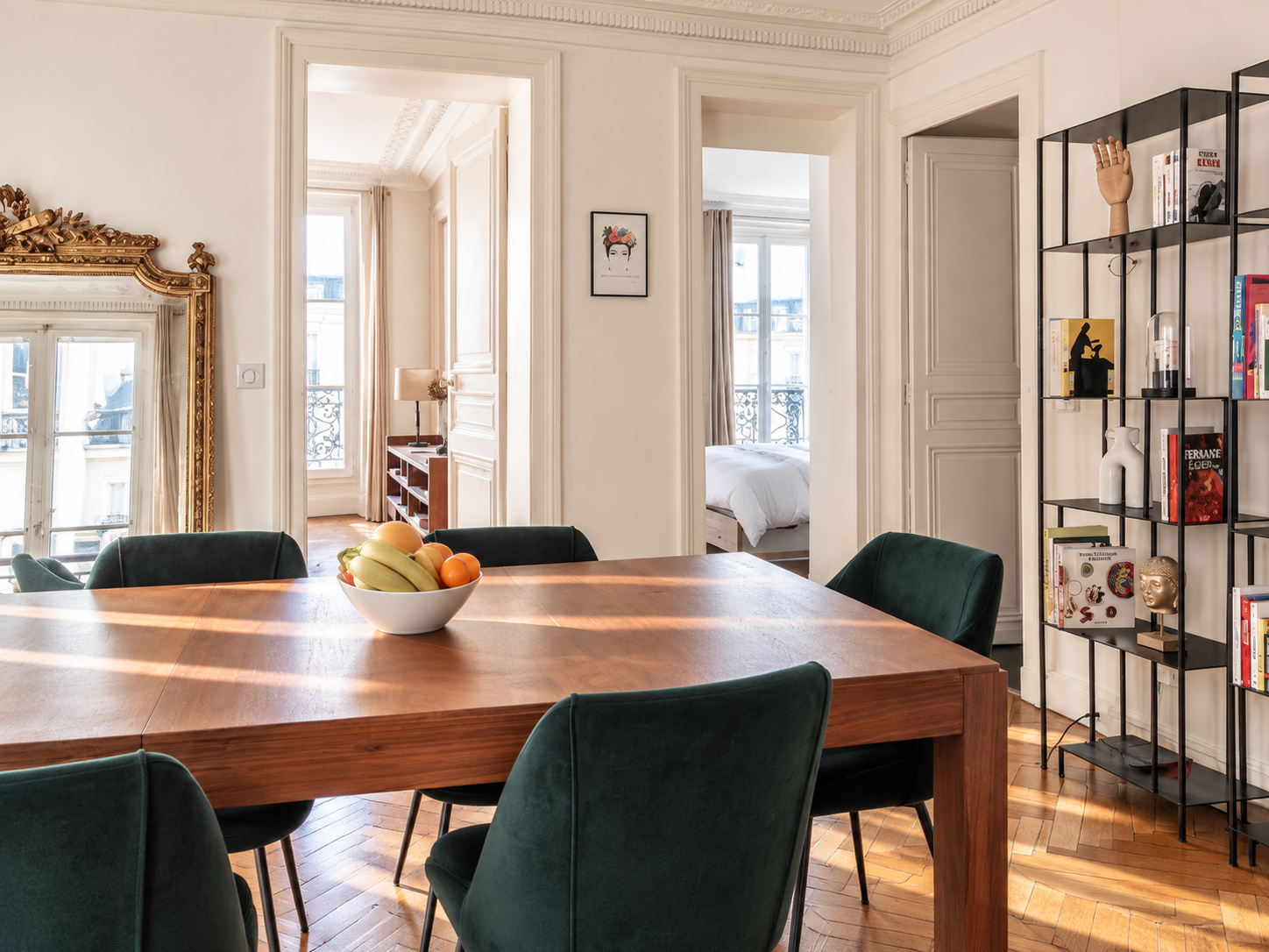 A dining room with a wooden table and six green velvet chairs, a bowl of assorted fruits, and a black metal bookshelf with decorative items and books. There are doorways leading to a bedroom and another room with windows and a balcony visible.