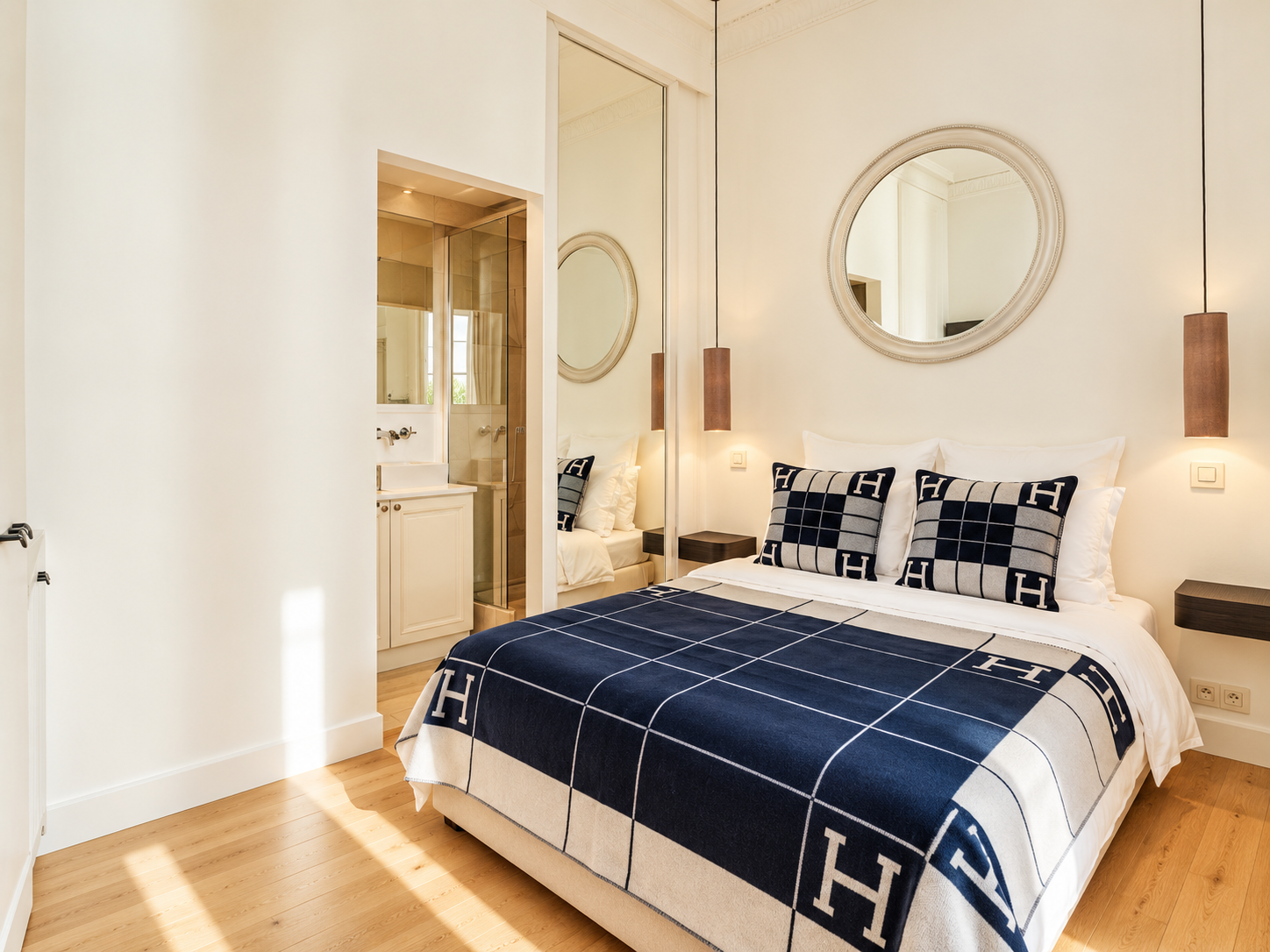 A bedroom with a bed covered in a navy blue and beige H-patterned blanket, two matching pillows, and a small mirror above the headboard. There is a small bedside table on each side of the bed, with pendant lights hanging from the ceiling. A door opens into a bathroom with a shower and a small sink. The room has wooden flooring and minimalistic decor.