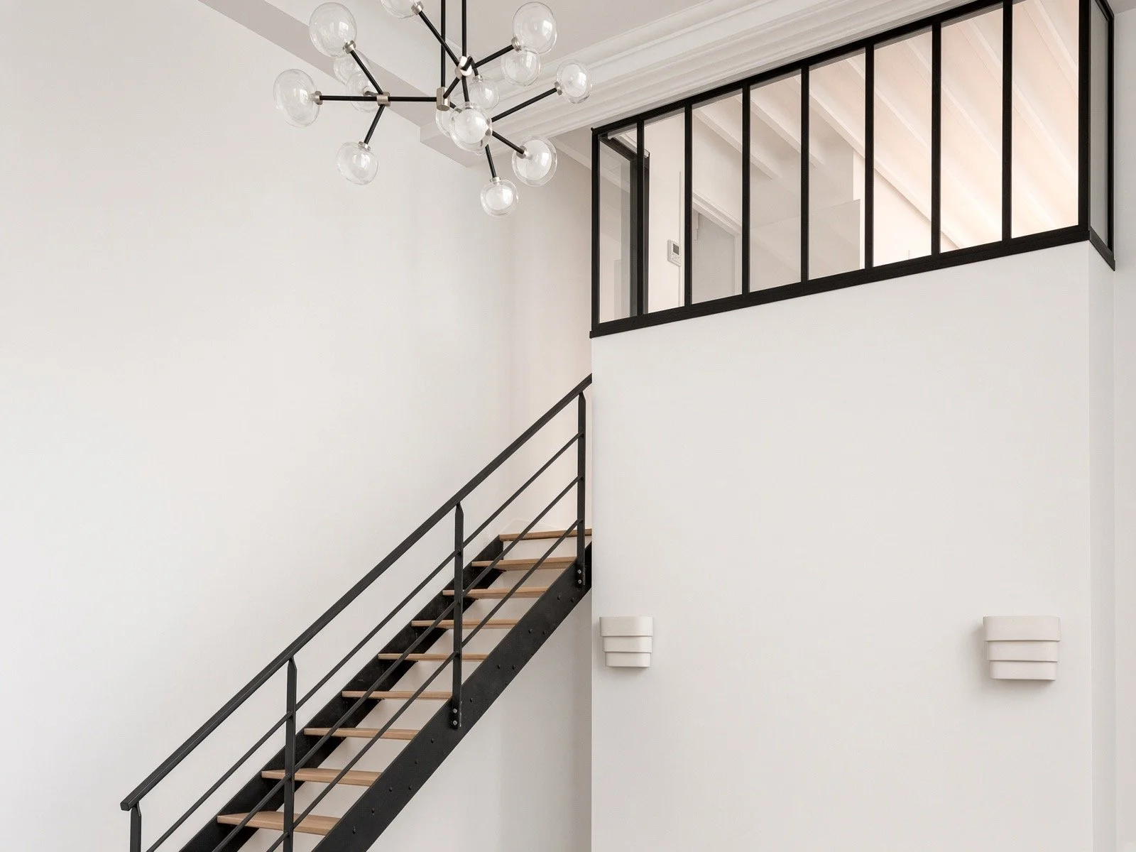 Modern staircase with black railing and wooden steps, white wall, contemporary chandelier with multiple exposed bulbs, black framed glass balcony, minimalist wall sconces.