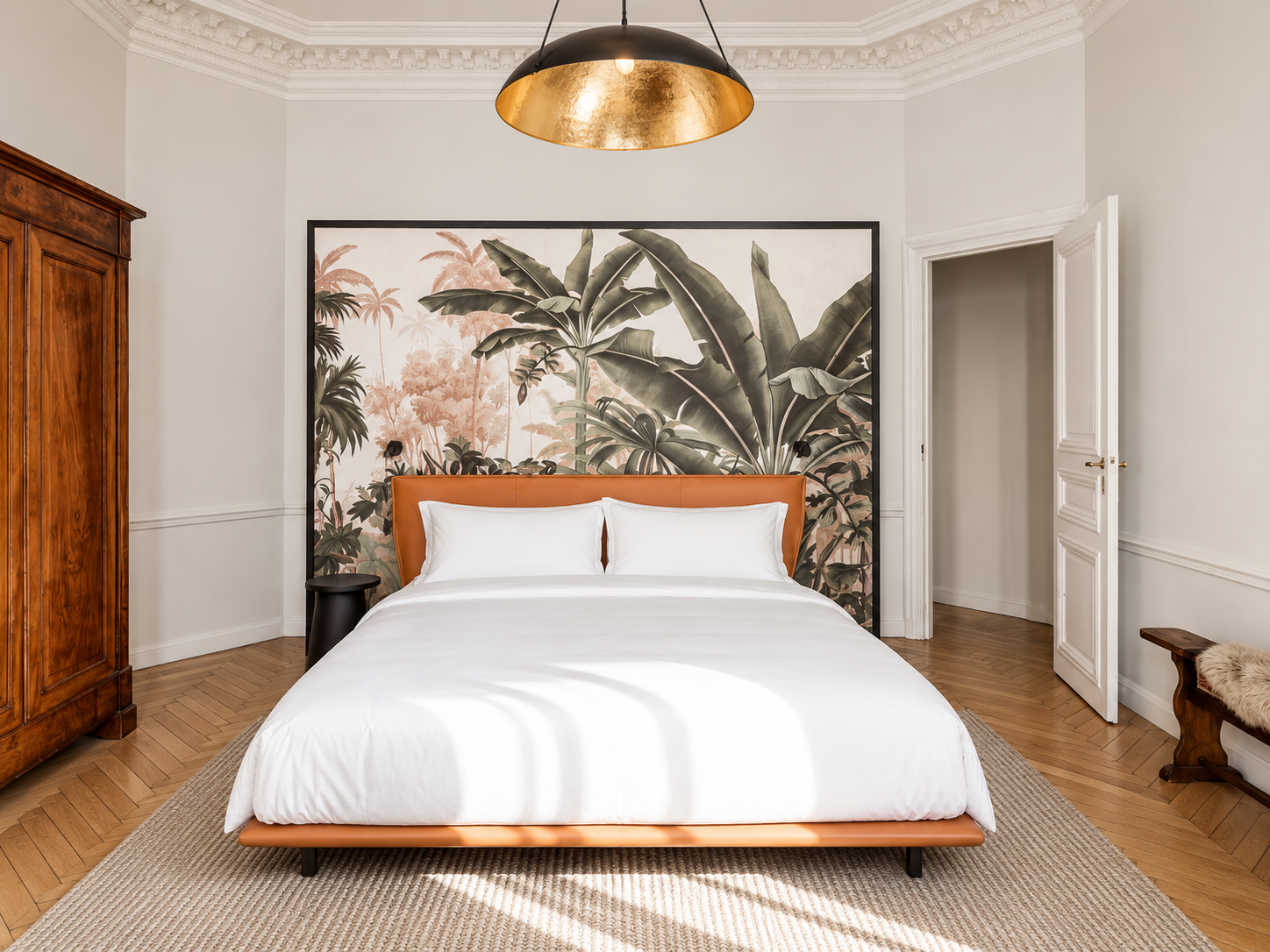 A bedroom with a modern bed featuring a tan leather headboard and white bedding. Behind the bed, there is a large tropical leaf mural. To the left, there is a wooden wardrobe, and to the right, a white door. The room has a beige rug and wooden flooring, with a black and gold ceiling lamp hanging above.