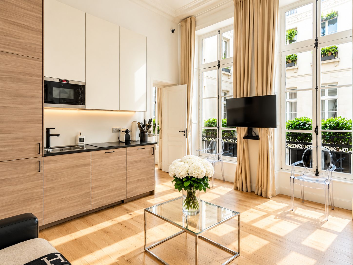 Bright living room with wooden cabinetry, a small kitchenette, a glass coffee table with a white flower arrangement, large windows with beige curtains, a wall-mounted TV, and transparent chairs.