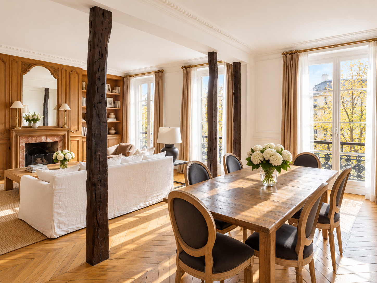 A bright living and dining room with large windows, white walls, wooden beams, a white sofa, a wooden dining table with six chairs, a vase of white flowers, a fireplace with a mirror and lamps, and built-in bookshelves.