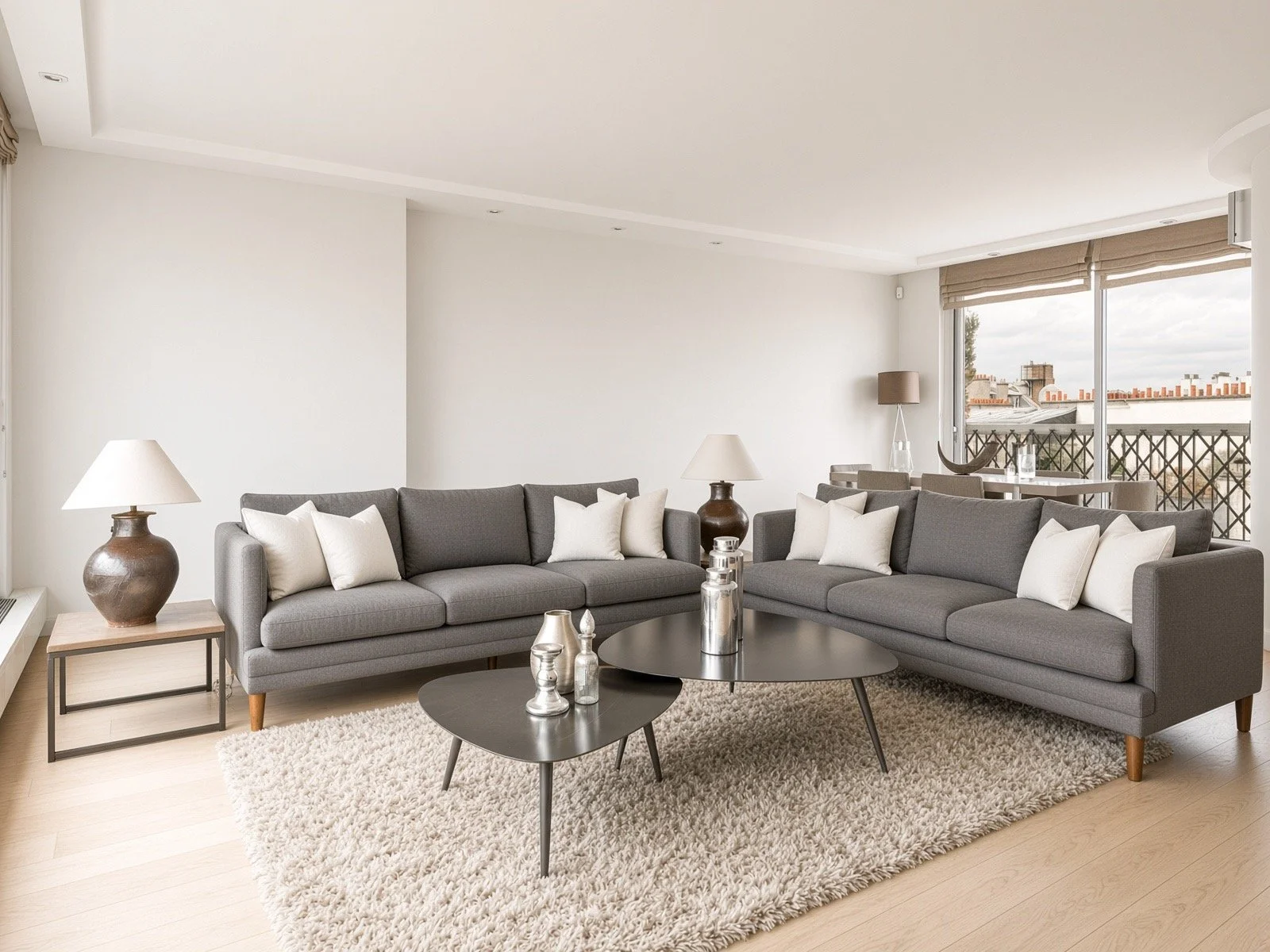 Modern living room with two gray sofas, white cushions, two black coffee tables, table lamps, a beige rug, and large windows with view of city rooftops.