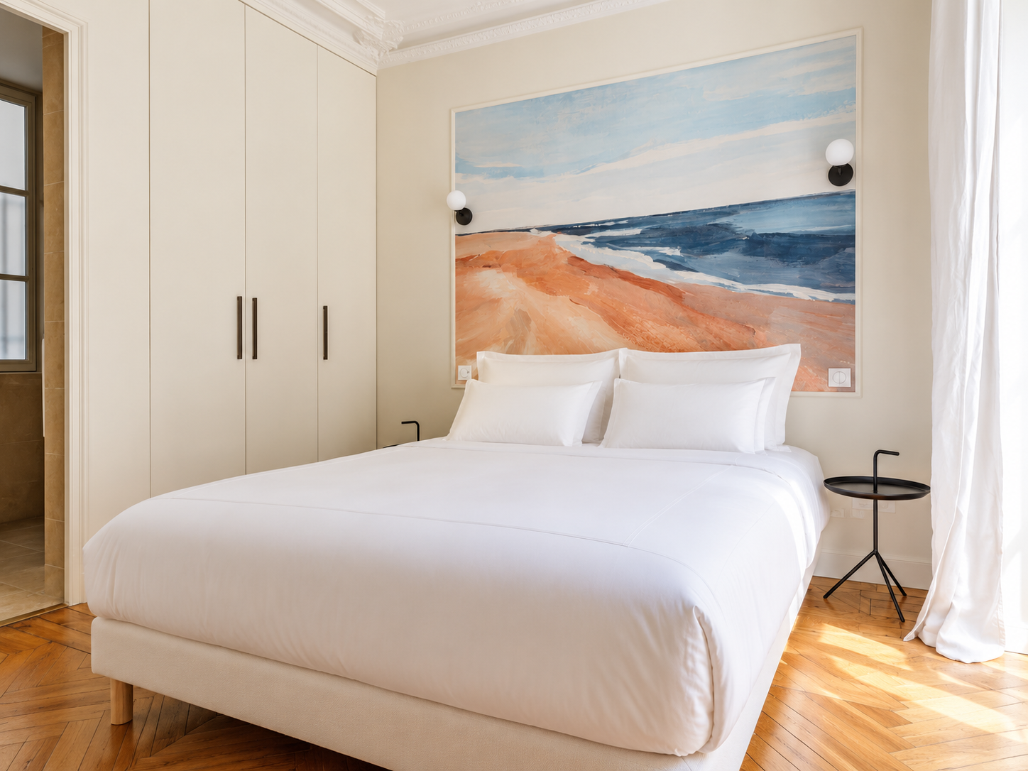 A bedroom with a white bed, large beach-themed painting behind it, and minimalist black side table with a straw. White curtains let sunlight into the room with hardwood floors.