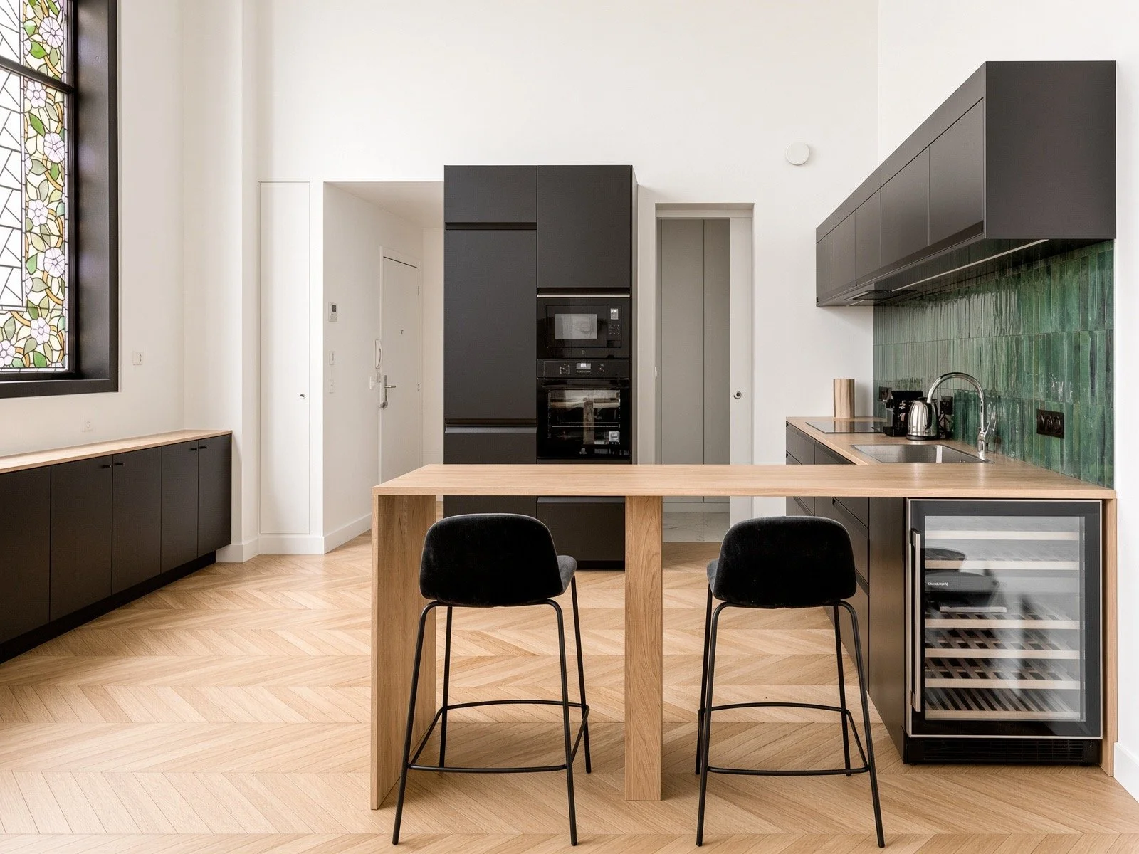 Modern kitchen with black cabinets, a wooden dining table, black chairs, green tiled backsplash, built-in appliances, and a wine fridge.