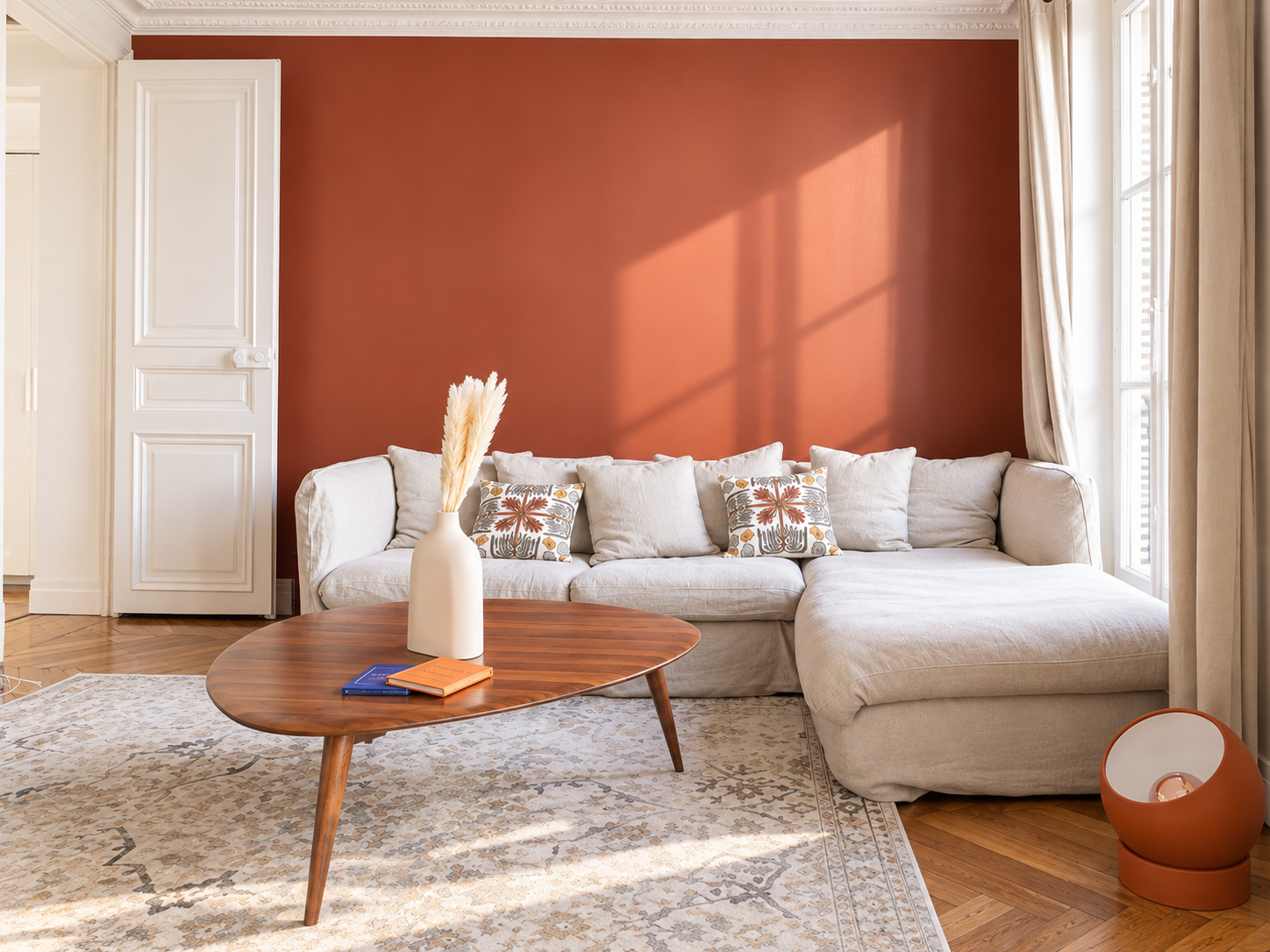 Living room with a large white sectional sofa, decorative pillows, a wooden coffee table with a vase of dried plants, a colorful rug, a large window with white curtains, and a terracotta-colored accent wall.