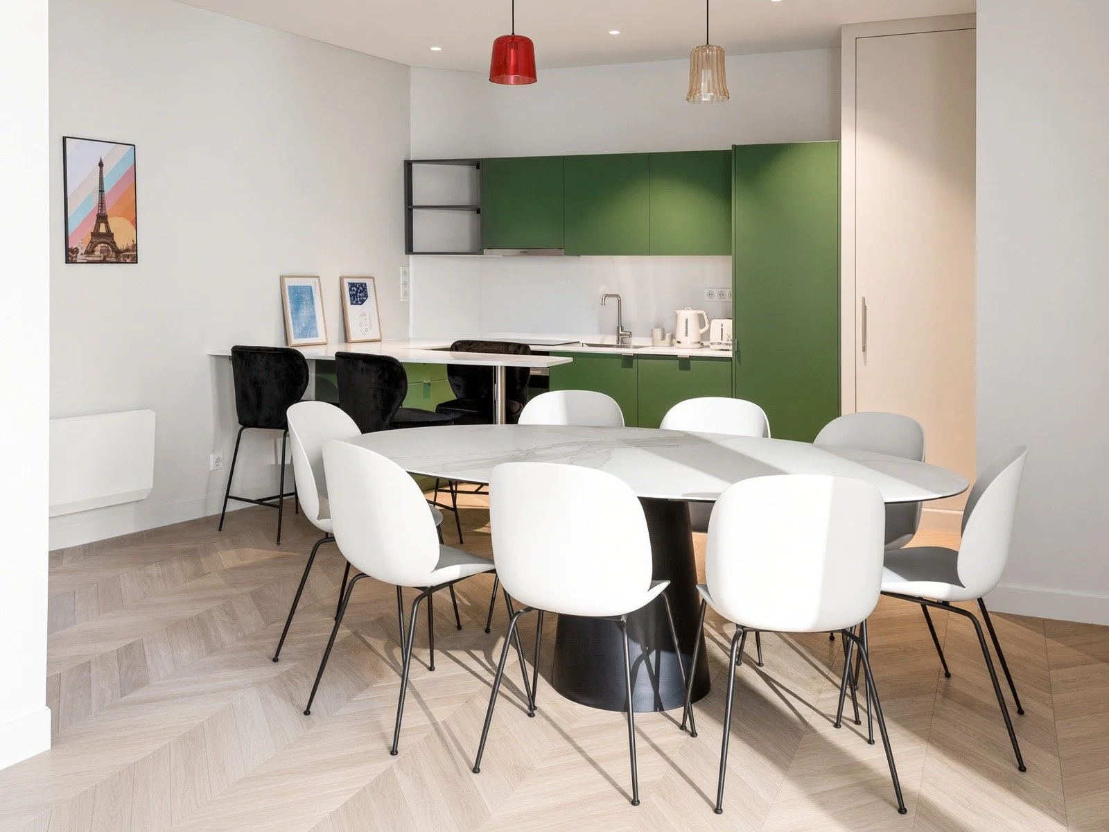 A modern dining area with a round marble table surrounded by white chairs. In the background, a kitchen with green cabinets, a white countertop, a sink, and appliances. The walls are white, and there are three hanging pendant lights in red, beige, and clear glass. There's framed artwork on the wall and a light wood herringbone floor.