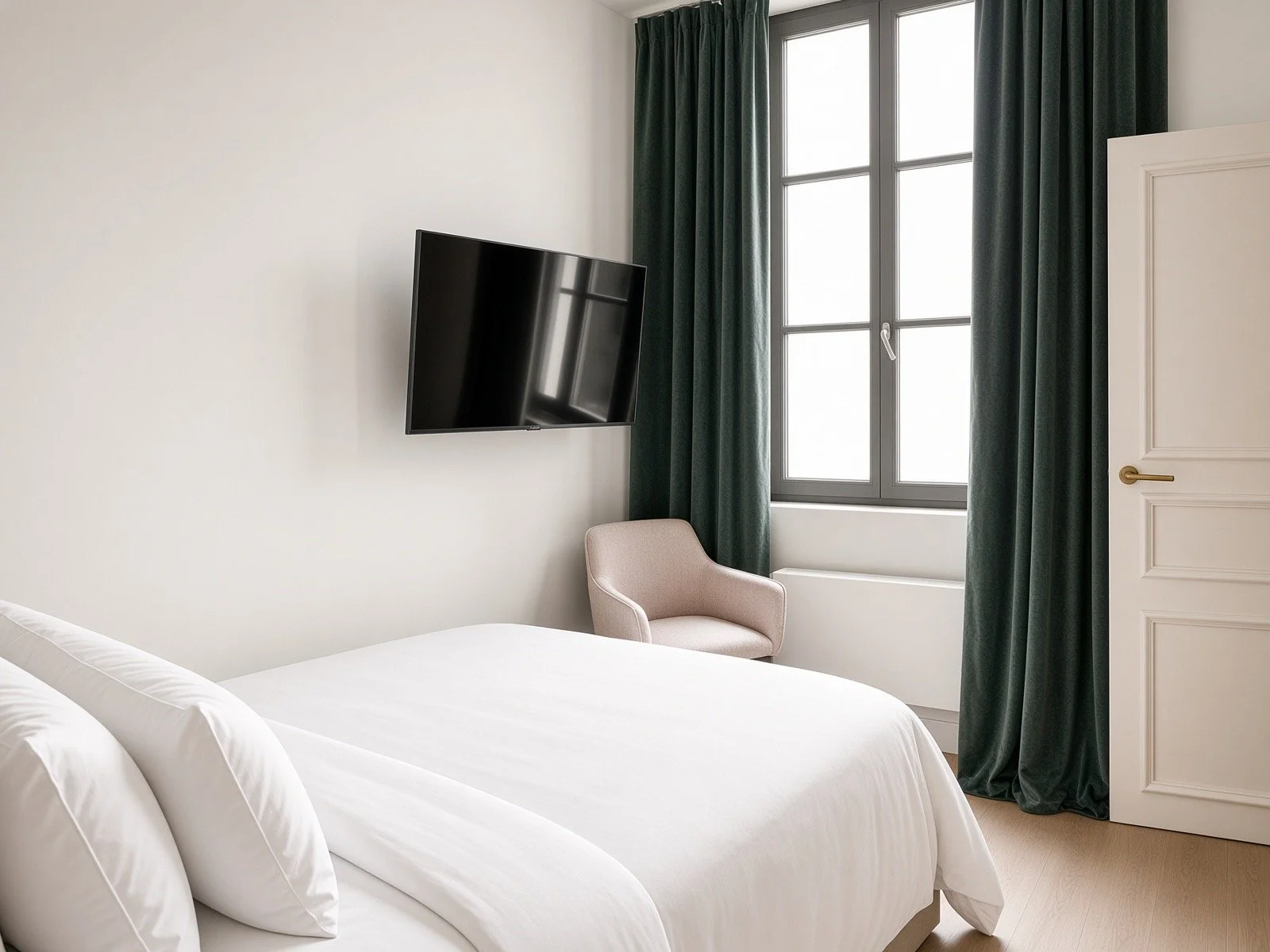 A bedroom with a bed, a wall-mounted TV, a beige armchair, a window with dark green curtains, and a white door.