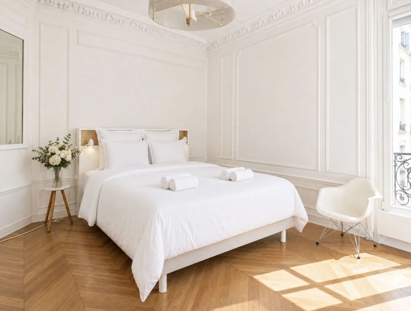 Bright, modern bedroom with white walls, a large white bed with four pillows, three rolled towels on the bed, a small side table with a vase of white flowers, a white chair near a window, and a ceiling light fixture.