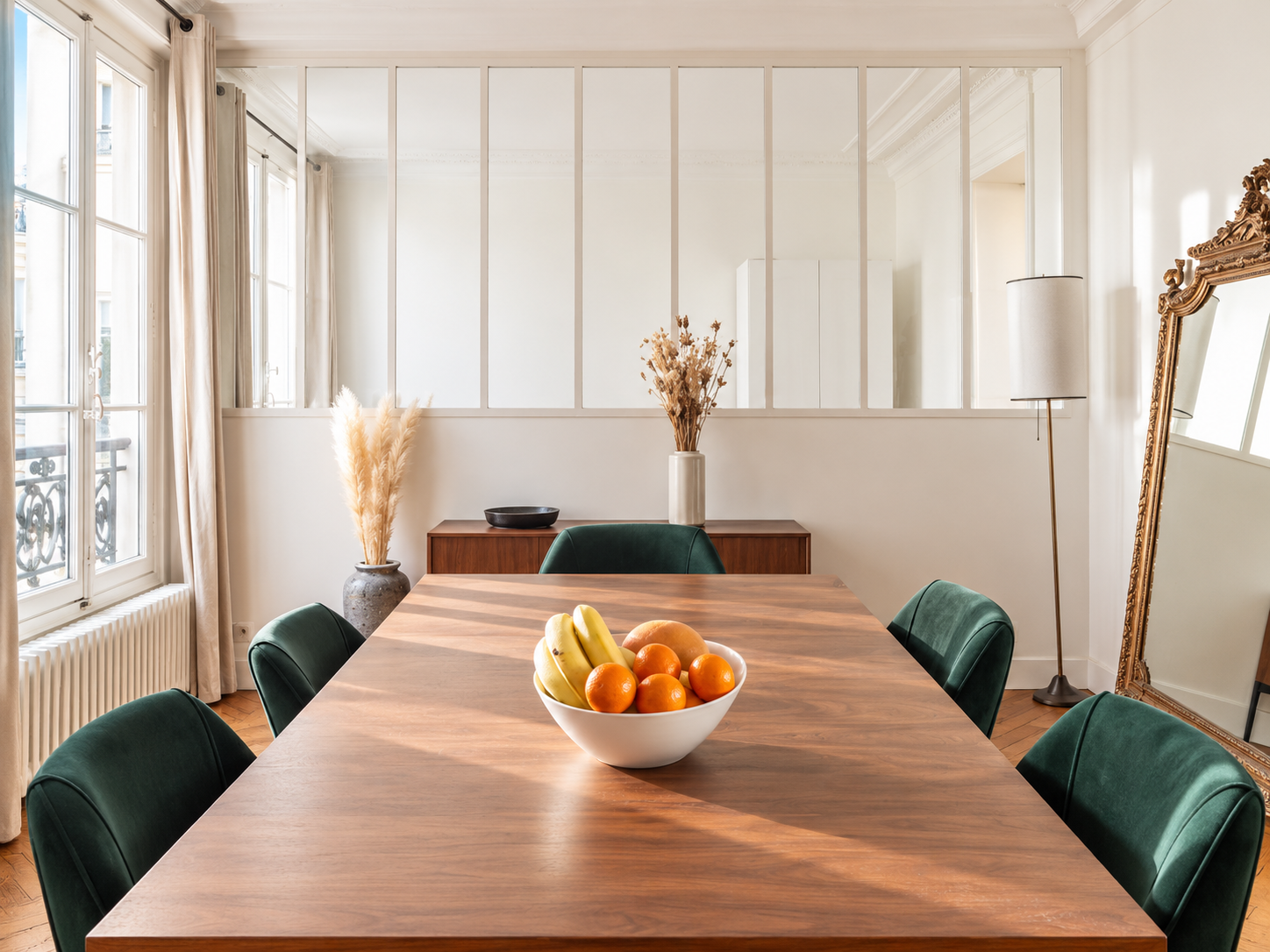 Modern dining room with a large wooden table, green chairs, a white fruit bowl with bananas and oranges, beige curtains, and a tall mirror with a gold frame.