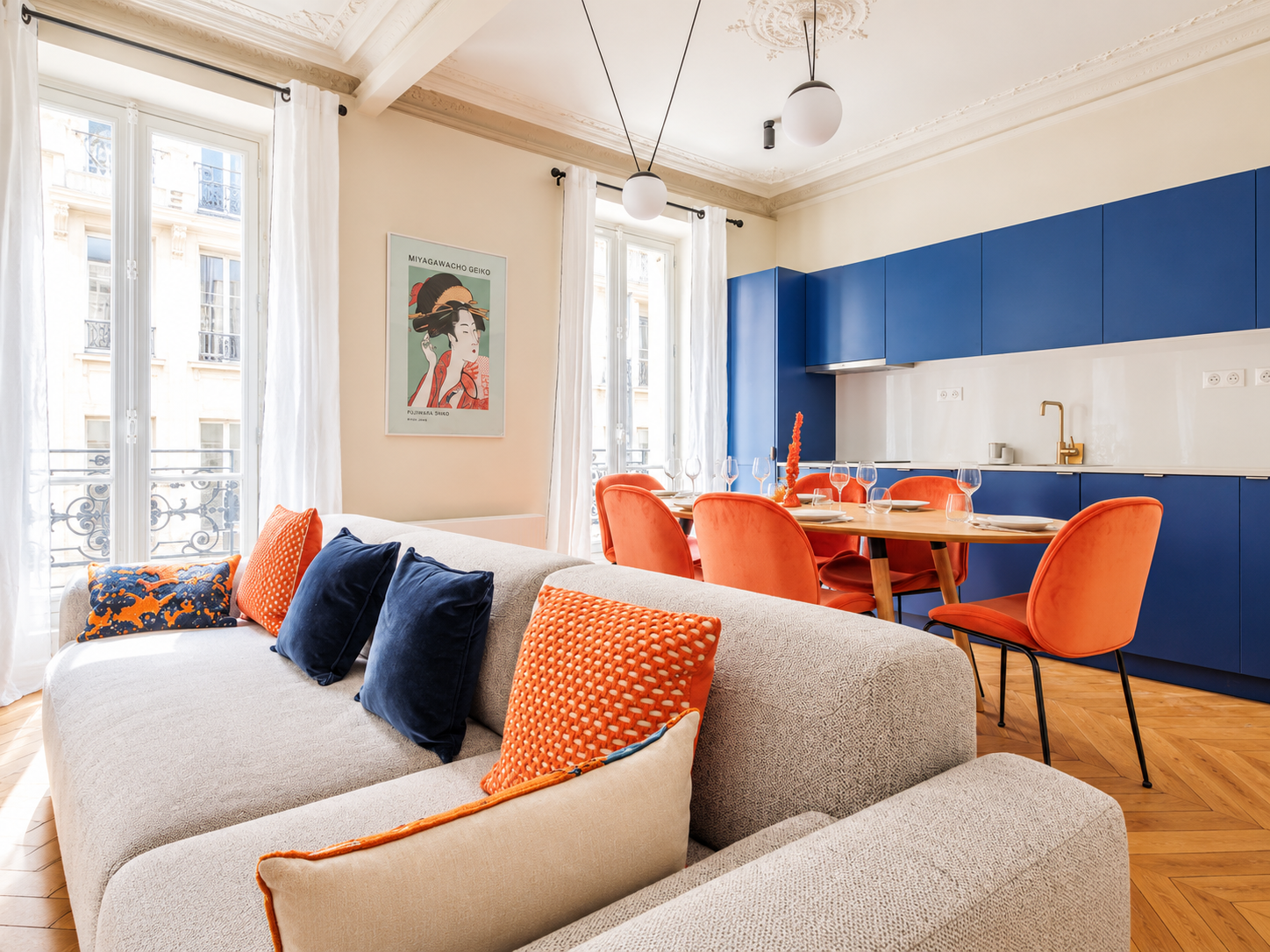 Living room and dining area with a beige sofa, colorful pillows, orange dining chairs, blue kitchen cabinets, a wooden dining table set for a meal, and large windows with white curtains.