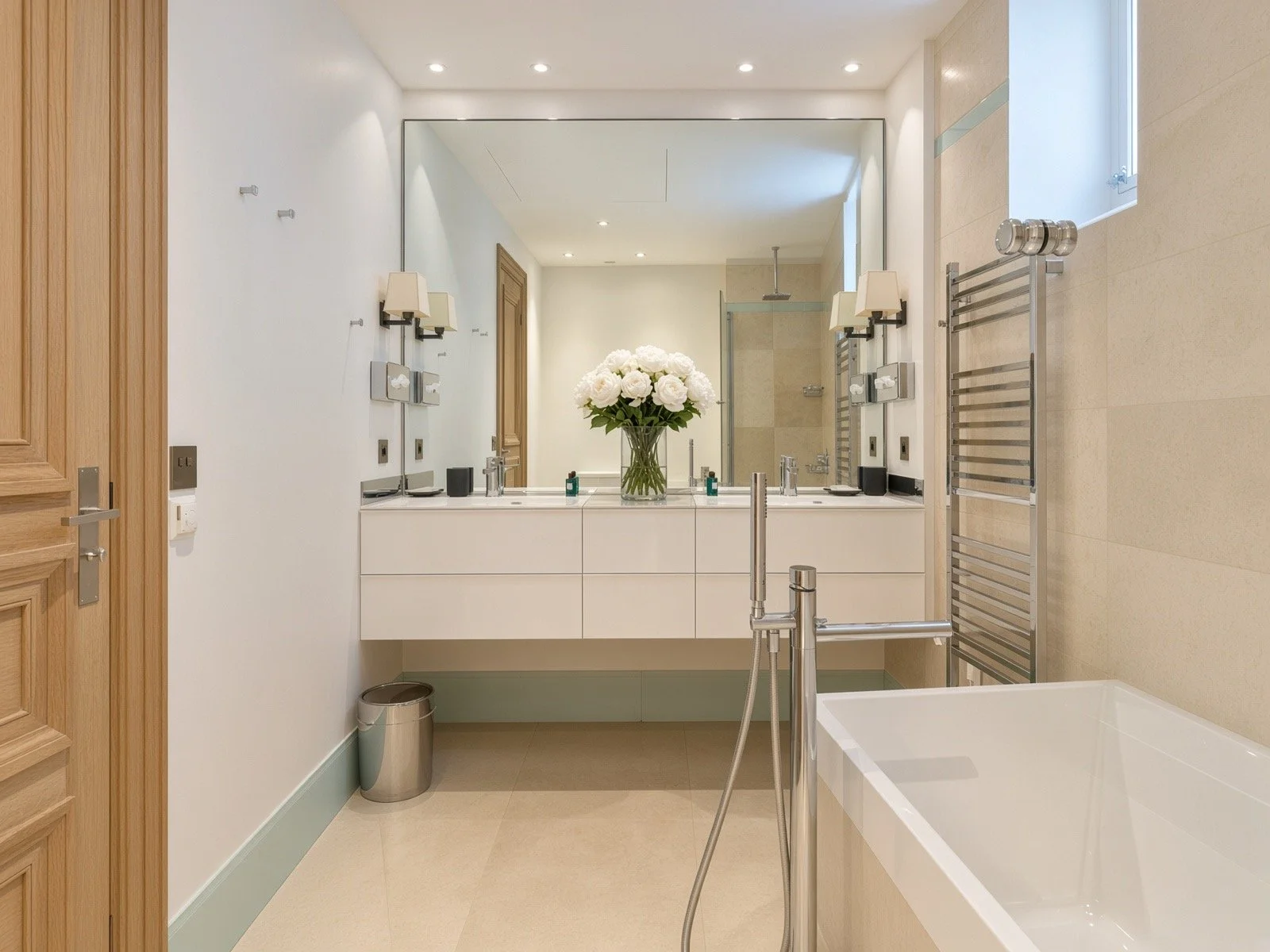 A modern bathroom with a large mirror, white vanity, a vase of white roses, a bathtub, a towel warmer, and a walk-in shower with glass enclosure.