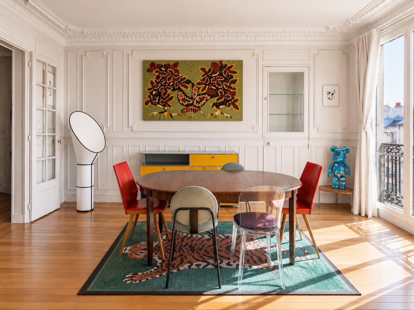 A dining room with a round wooden table surrounded by six chairs of various styles and colors. The room has a large window with curtains, a green and orange tiger-themed rug, white paneled walls, and artwork on the walls. There is a yellow and grey sideboard, a white floor lamp, and a blue cardboard bear figure on a small table by the window.