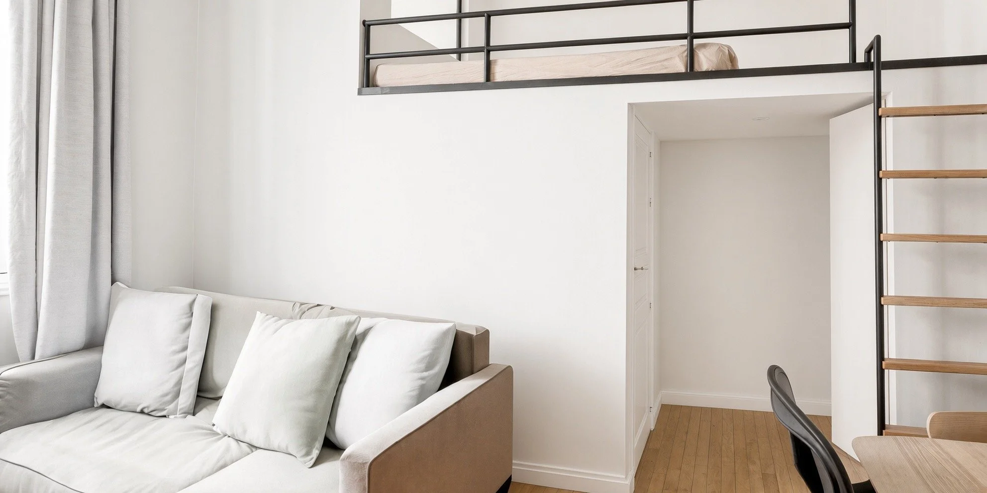 Interior of a modern apartment with a white sofa and pillows, a lofted bedroom with a black metal railing, and a small open hallway with a wooden ladder.