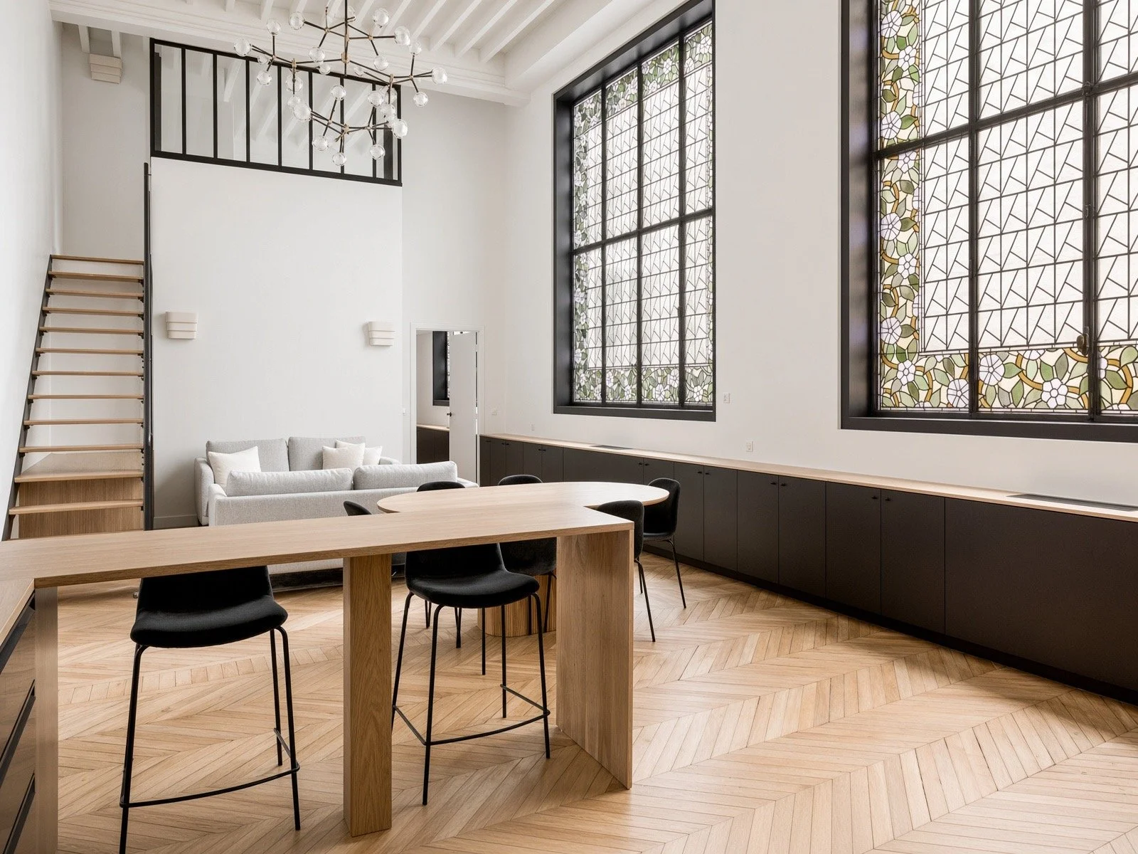 Interior of a modern, minimalistic room with large stained glass windows, a contemporary chandelier, a gray sofa, a wooden table with black chairs, and built-in black storage cabinets along the wall.