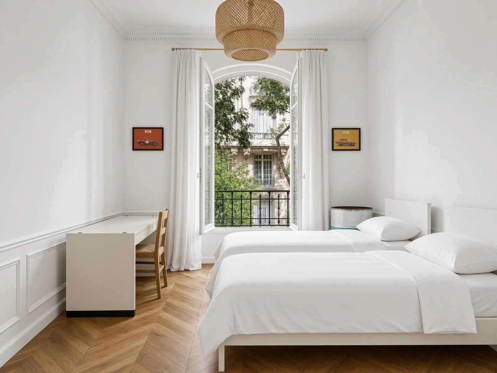 A bright hotel room with two white beds, a nightstand, and a large arched window with white curtains, overlooking a city street lined with trees.