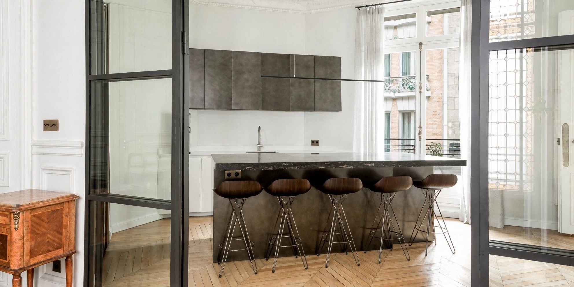 Modern kitchen area with a dark gray island, bar stools, white cabinetry, a gray backsplash, and large windows with view of neighboring buildings.