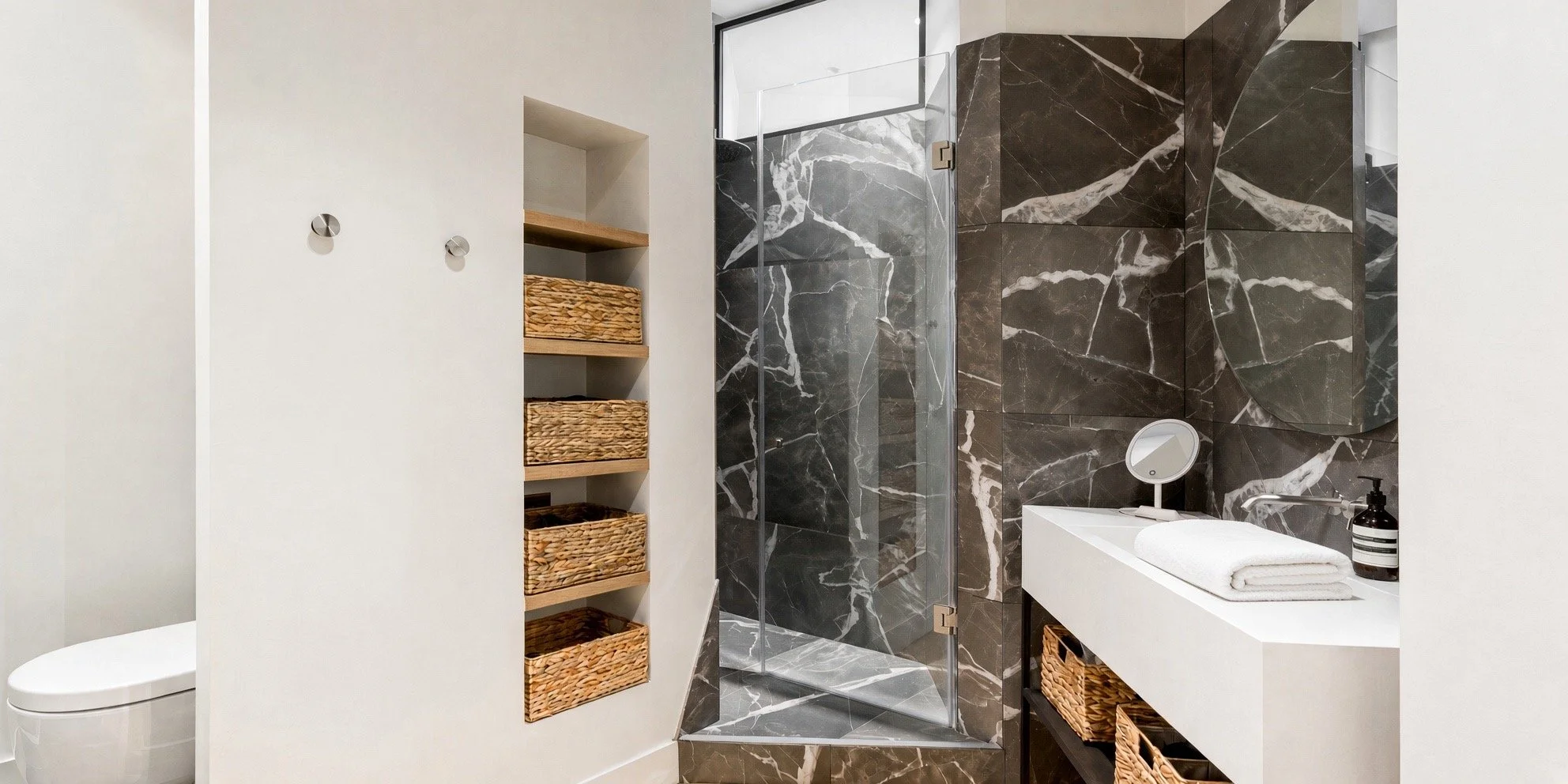 Modern bathroom with white walls, black marble accent walls, a glass shower enclosure, a white sink with a mirror, wicker baskets on a black shelf, and a black soap dispenser.