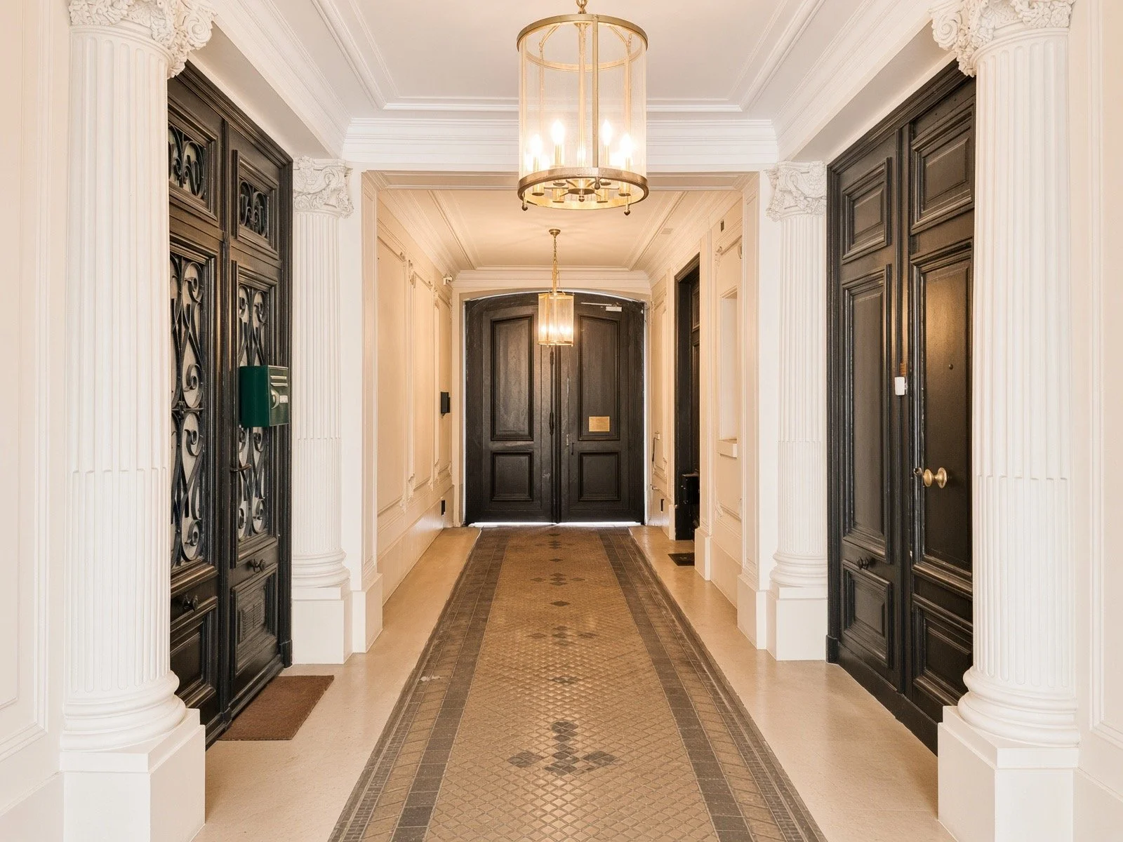 Interior hallway with white walls, ornate decorative columns, black front doors, chandelier lighting, and a patterned runner rug.