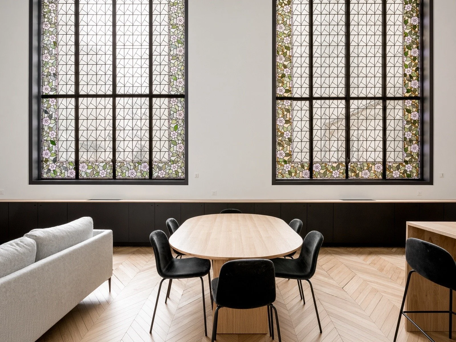 Interior of a modern room with two large stained glass windows featuring floral patterns, a wooden conference table surrounded by black chairs, a white sofa, and light-colored hardwood flooring.