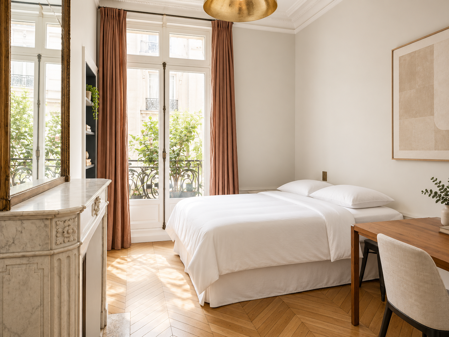 Bright room with a white bed, pink curtains, a wooden desk with a plant, and city view through French doors with black wrought iron railing.