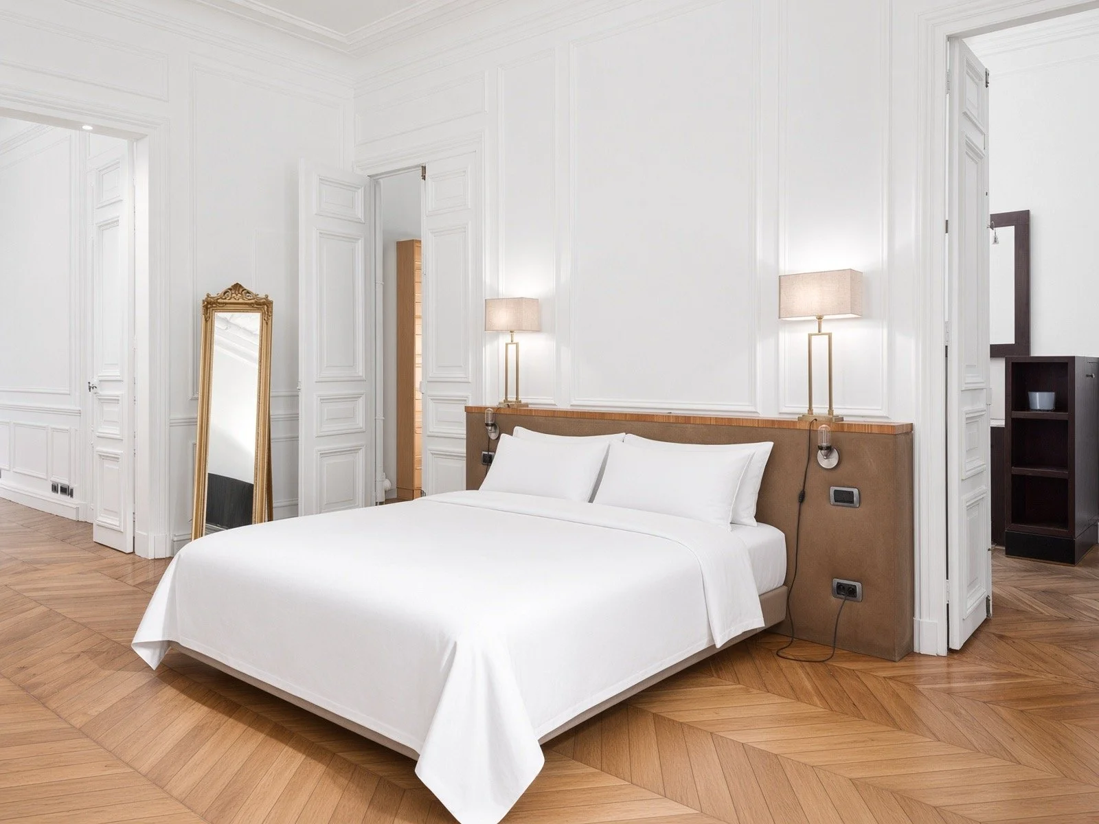Modern hotel bedroom with white walls, wooden floors, a large bed with white bedding, two bedside lamps, a tall mirror with a gold frame, and a black shelving unit in the corner.