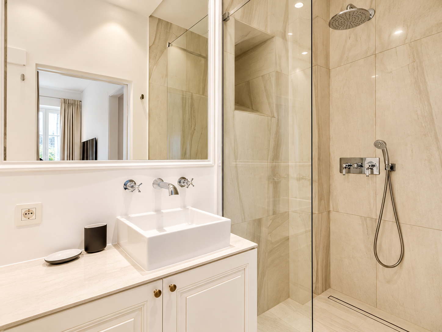 Modern bathroom featuring a large mirror, a white rectangular vessel sink with chrome faucets, beige marble tile shower area with a rainfall showerhead, and a handheld shower in the corner. The vanity has white cabinetry with brass knobs, and a small black cup and white dish are on the light-colored countertop.