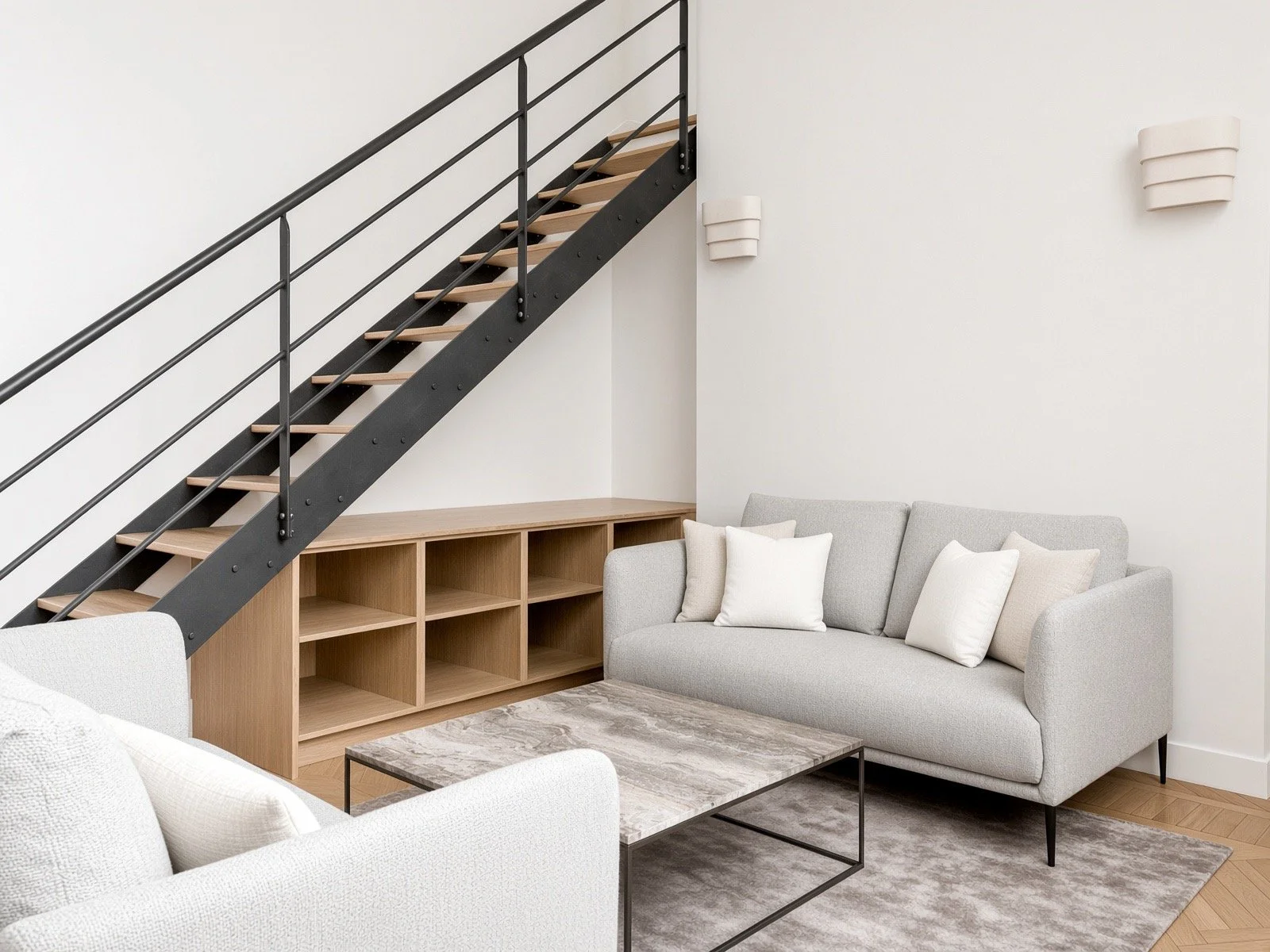 Living room with a gray sofa, white pillows, a wooden shelf, a marble coffee table, and a staircase with black metal railing against a white wall.