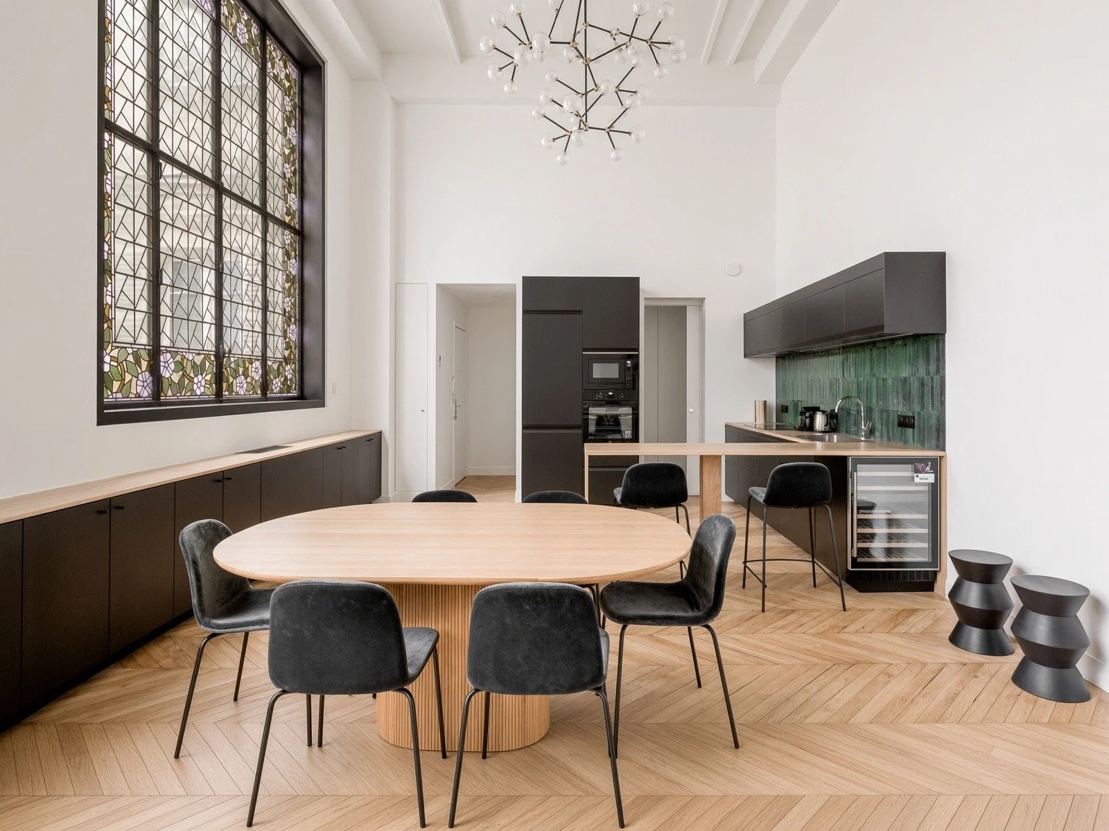 Modern kitchen and dining area with a round wooden table surrounded by black chairs, large window with stained glass, black cabinetry, green tiled backsplash, and decorative black stools.