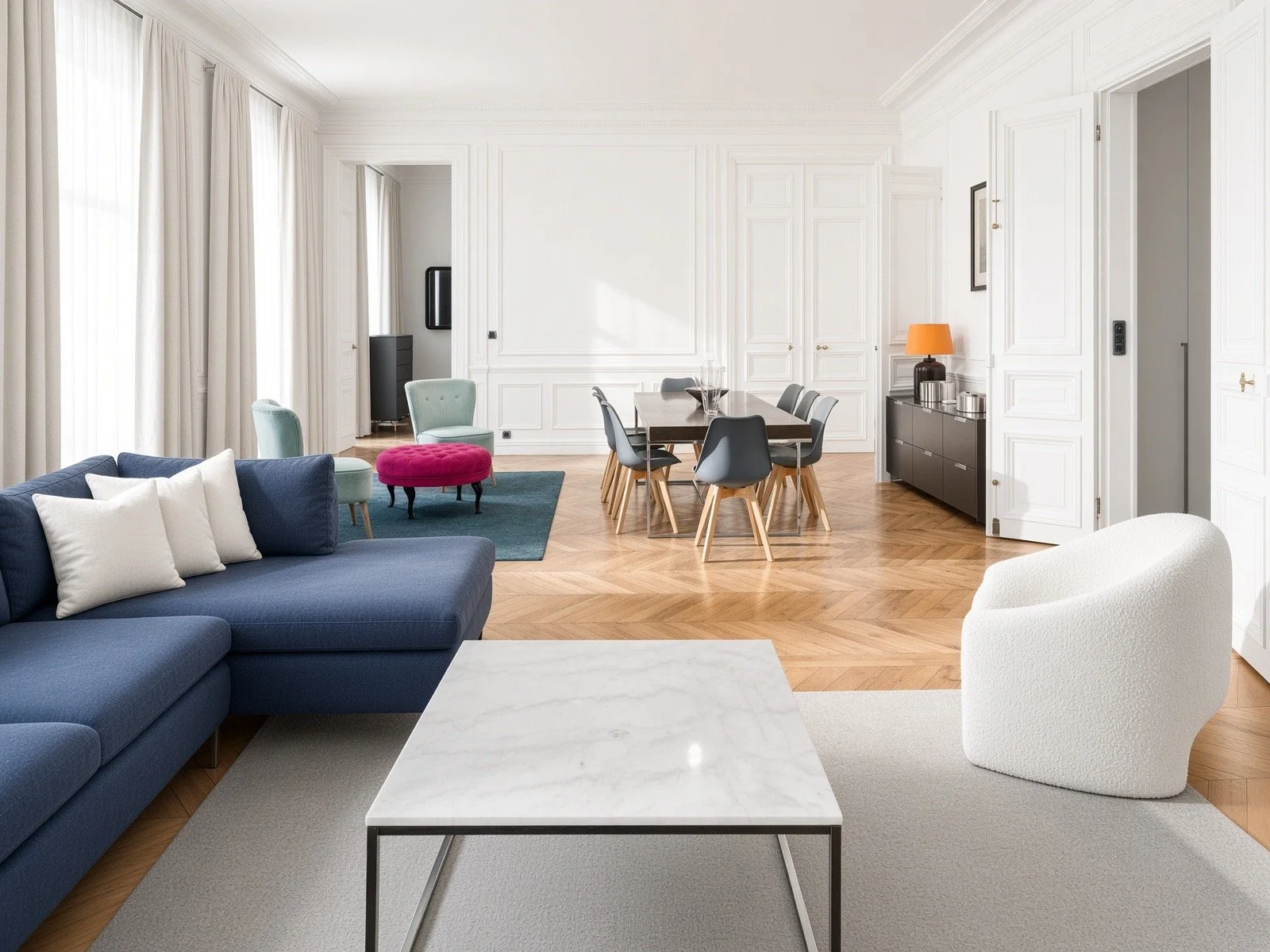 Bright, modern living room with white walls, large windows with curtains, a navy sofa with white pillows, a white textured armchair, a marble coffee table, a dark dining table with black chairs, and a black sideboard with a lamp and decor.