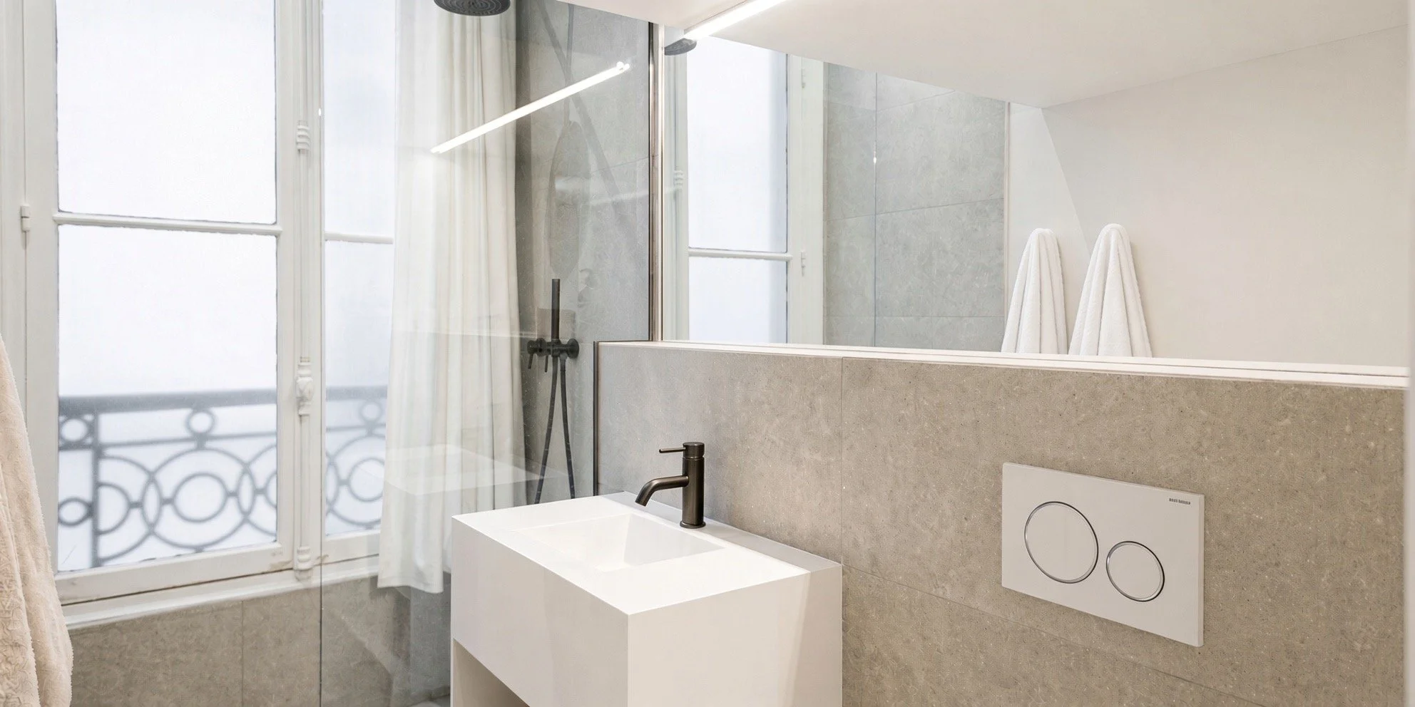 Modern bathroom with large window, white towels hanging, wall-mounted toilet buttons, small square sink with black faucet, and a walk-in shower with black fixtures.