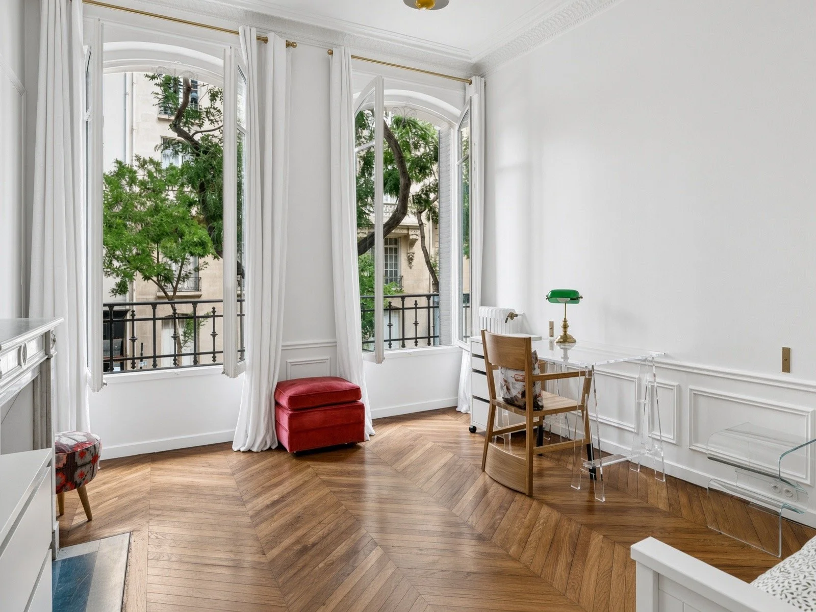 Room with large open windows, white walls, hardwood herringbone floor, small desk with green banker lamp, wooden chair, red ottoman, white curtains, and minimal decor.