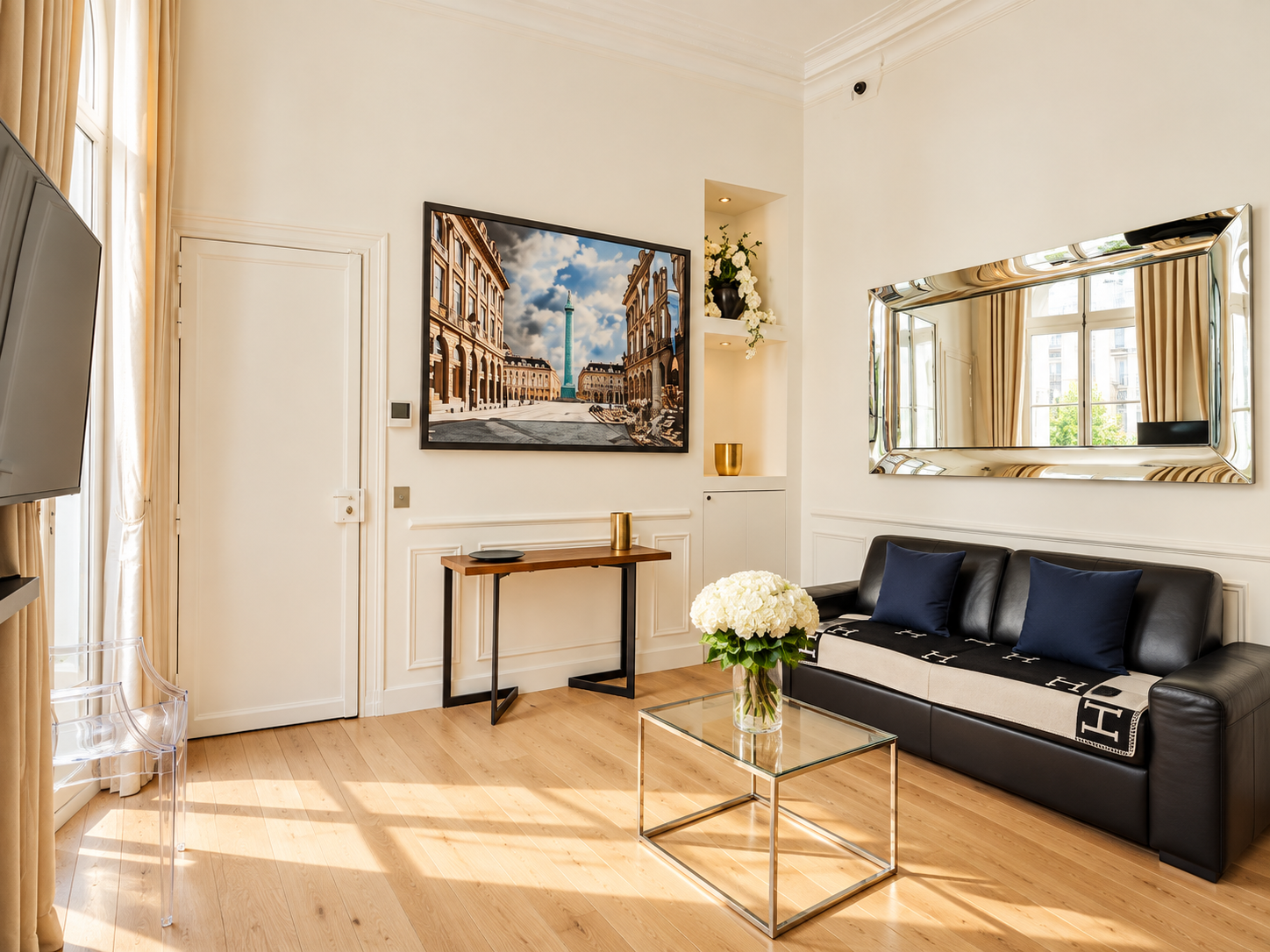 Elegant living room with a black leather sofa with navy pillows, a glass side table with white flowers, a wooden console table, a large framed photo of an architectural site, a sleek mirror, a wall nook with flowers, and a window with beige curtains.