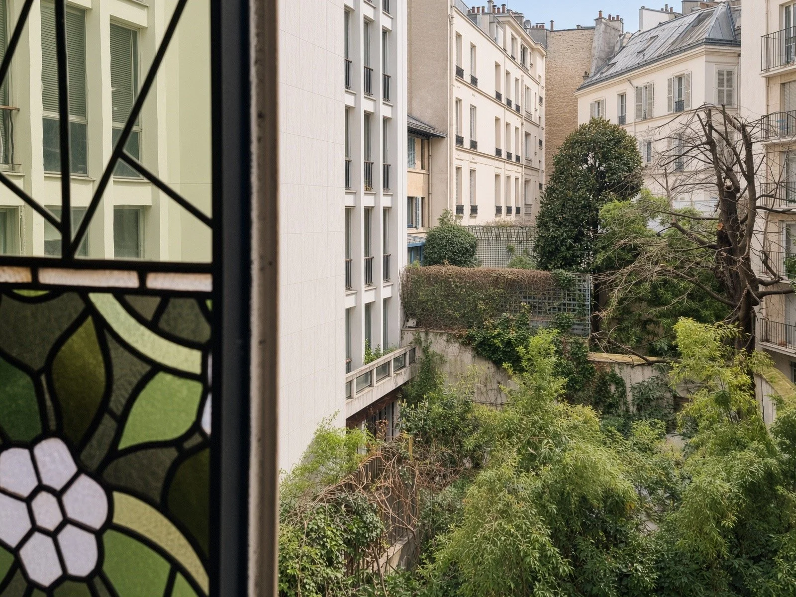 View of a city courtyard through a stained glass window with a black metal frame, showing buildings, trees, and greenery.
