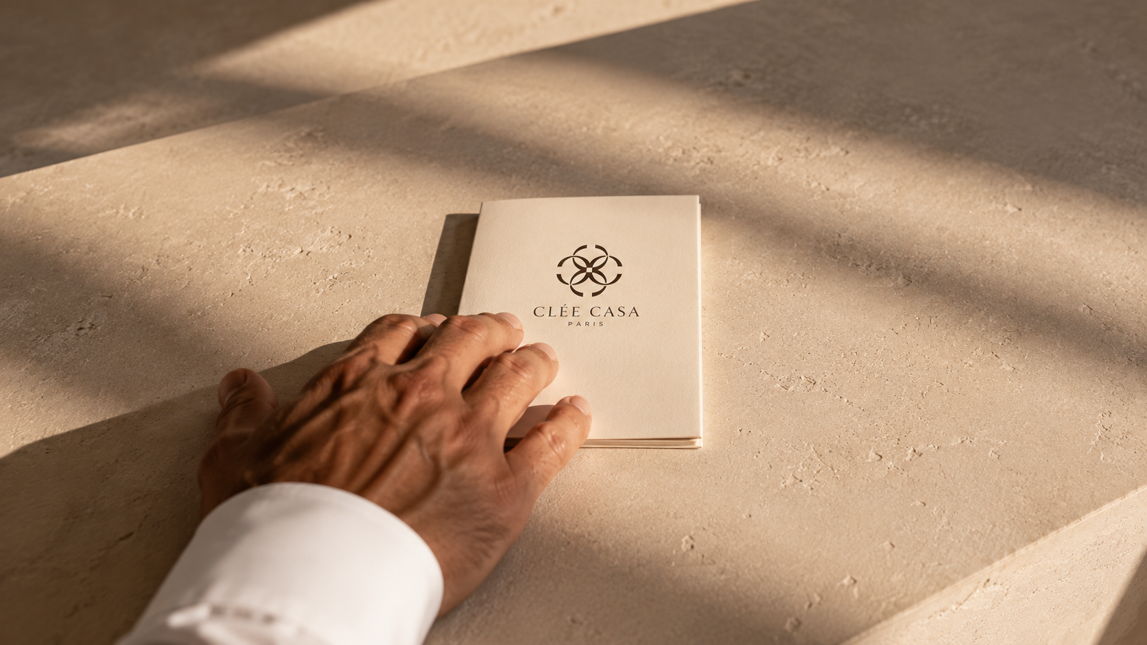 A hand resting on a cream-colored booklet with a geometric logo and the text 'Clée Casa Paris' on a beige stone surface, with shadow patterns from sunlight.