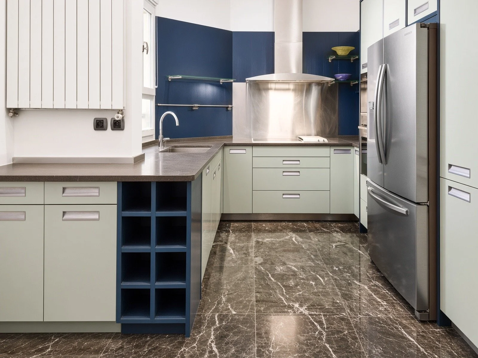 Modern kitchen with cream and blue cabinets, stainless steel refrigerator, marble floor, and a dark countertop. Blue paint accents the walls above the counter.