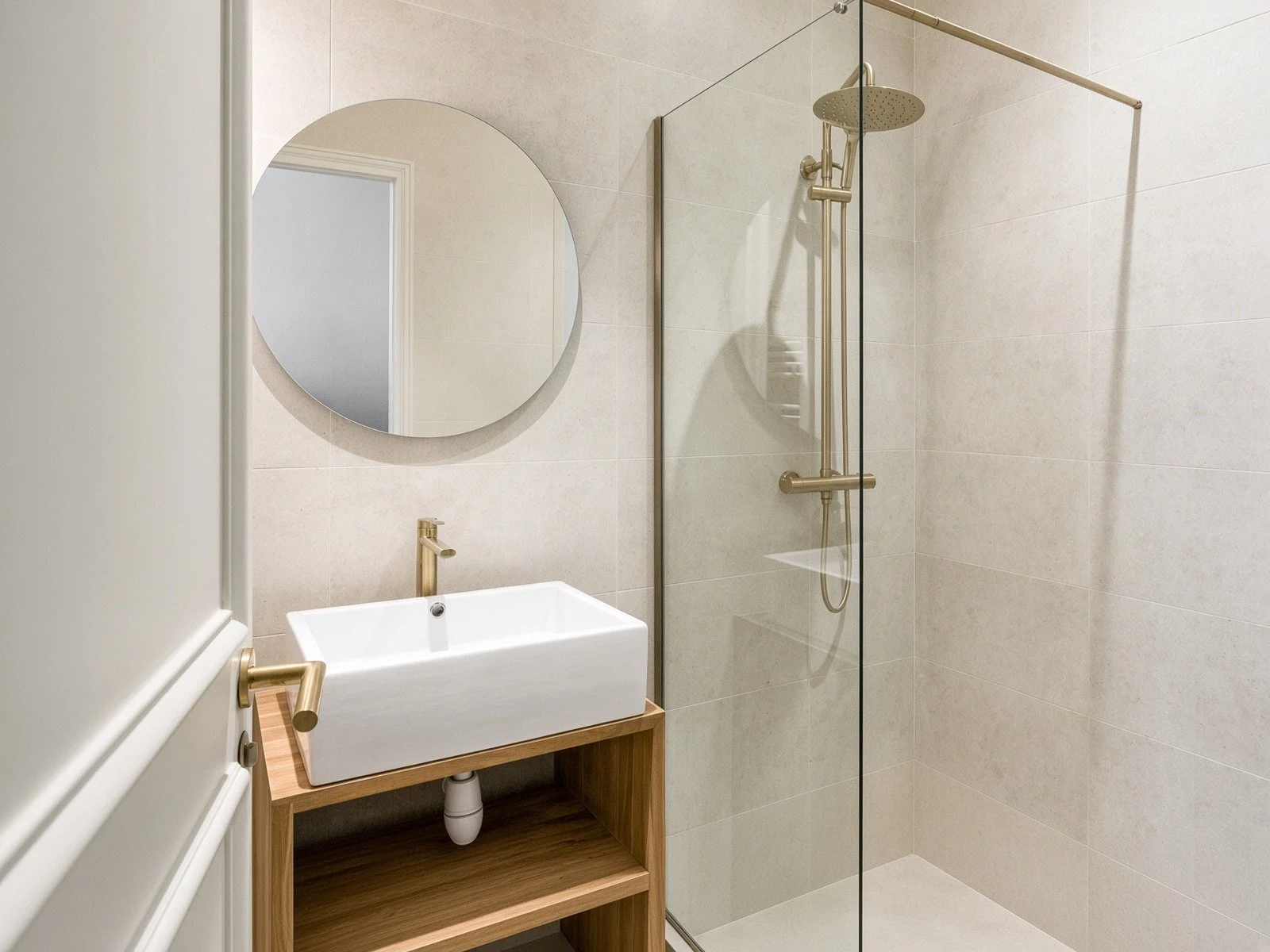 Modern minimalist bathroom with a rectangular vessel sink, oval mirror, glass-enclosed shower with gold fixtures, beige tiled walls, and a wooden vanity.