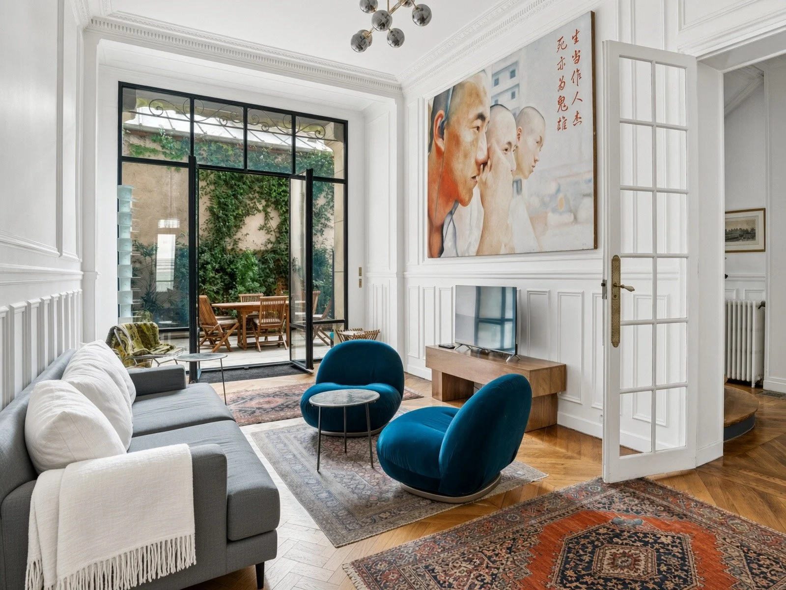 Living room with white paneled walls, a beige sofa with white and gray pillows, two blue velvet chairs, a wooden TV stand with a flat-screen TV, large artwork of three elderly Asian men, a glass door open to a patio with outdoor dining furniture, and a chandelier on the ceiling.