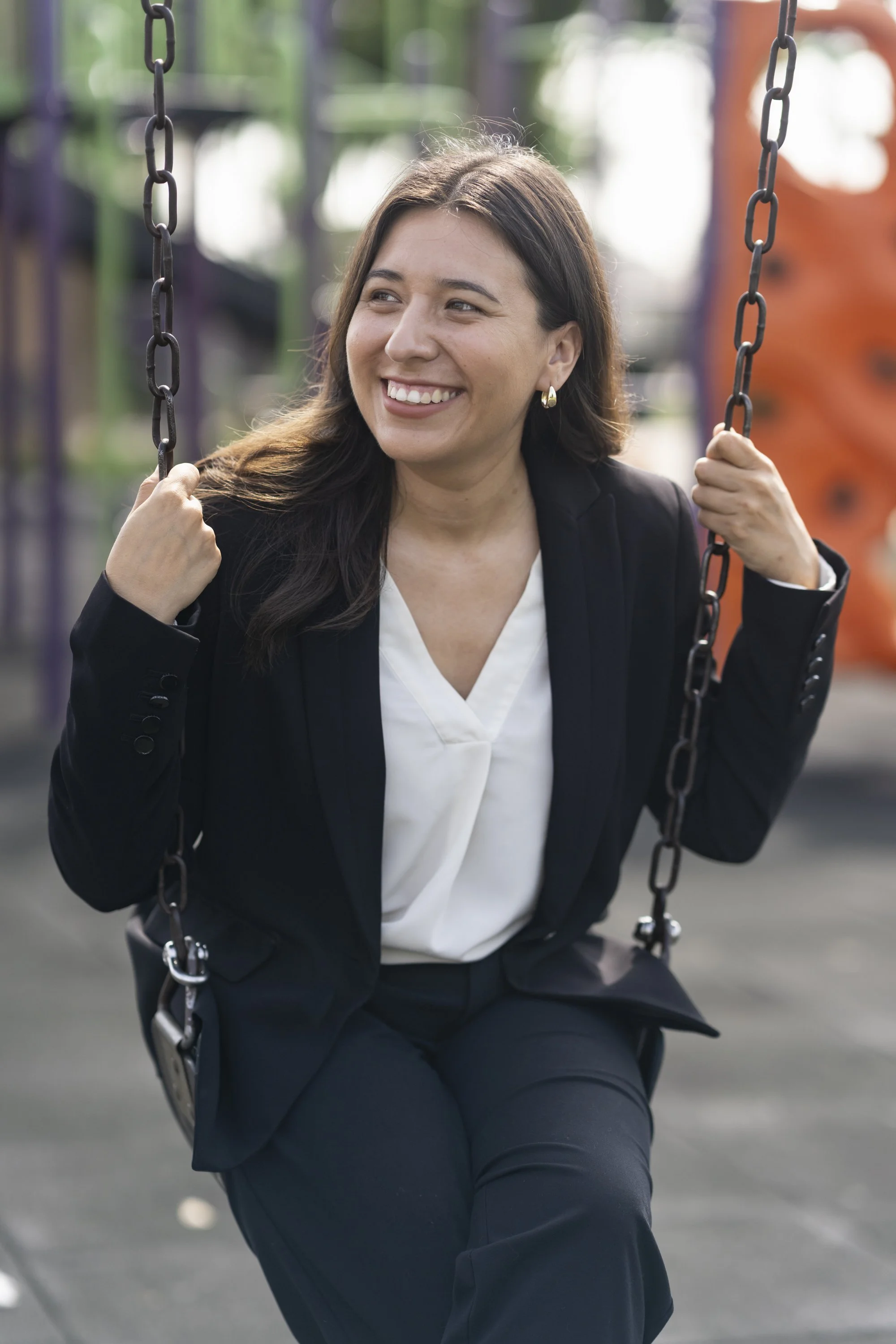 A woman in a black suit and white blouse sitting on a playground swing, smiling and holding the chains, with blurred colorful playground equipment in the background.