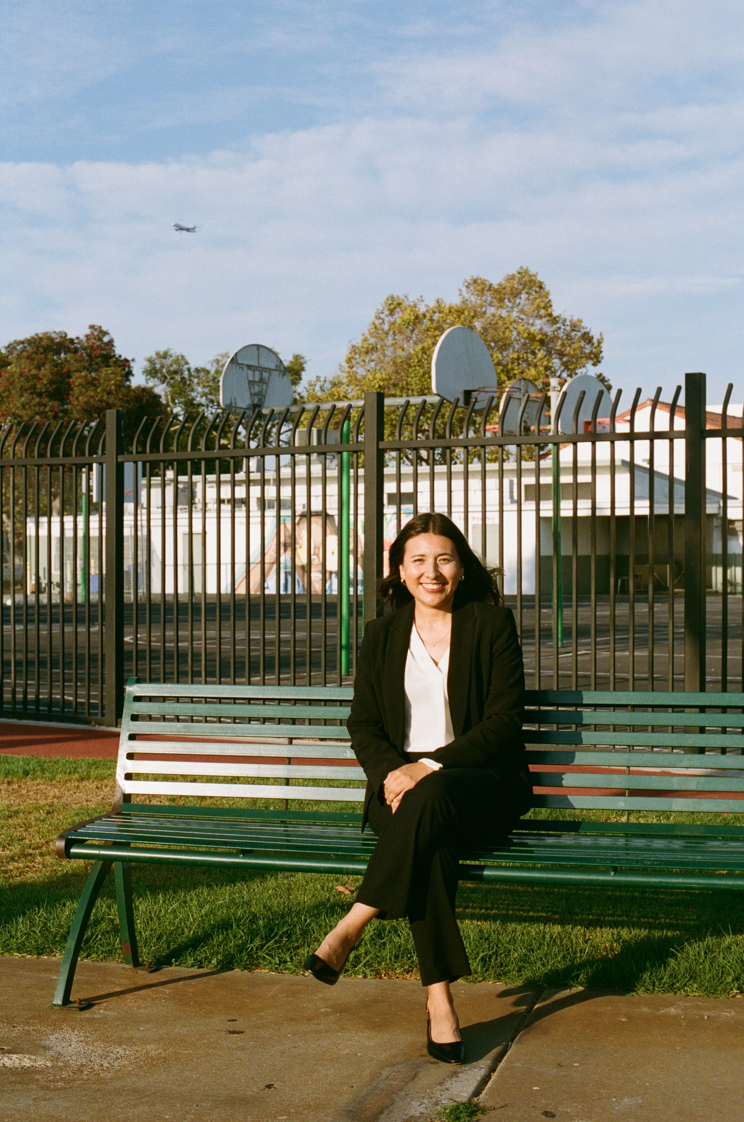 A woman in a black blazer and white blouse sitting on a green park bench outdoors, smiling, with a playground and trees in the background, and a small airplane flying in the sky.