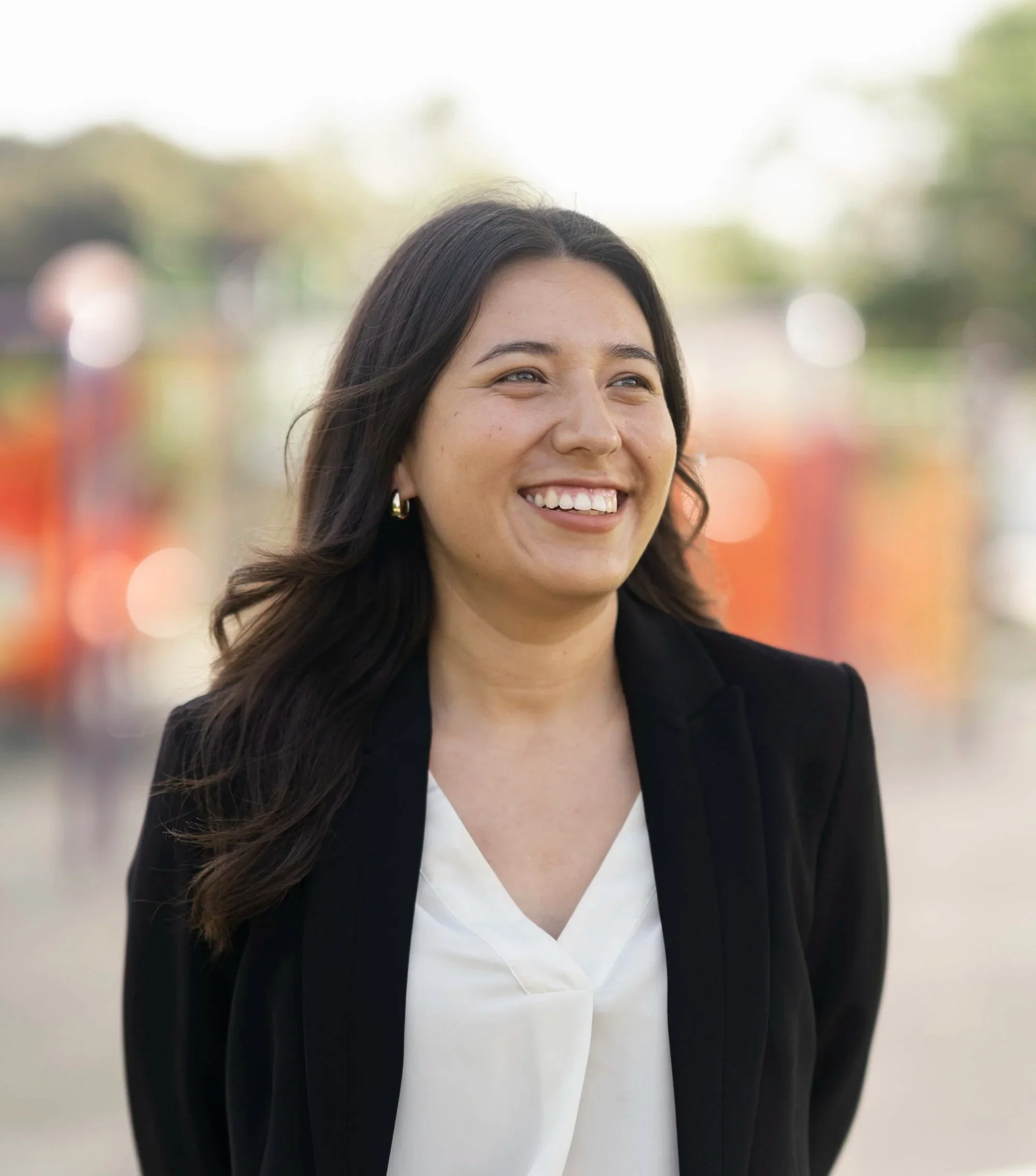 Close-up of a smiling woman with long dark hair, wearing earrings, dressed in professional attire, with a blurred outdoor background.