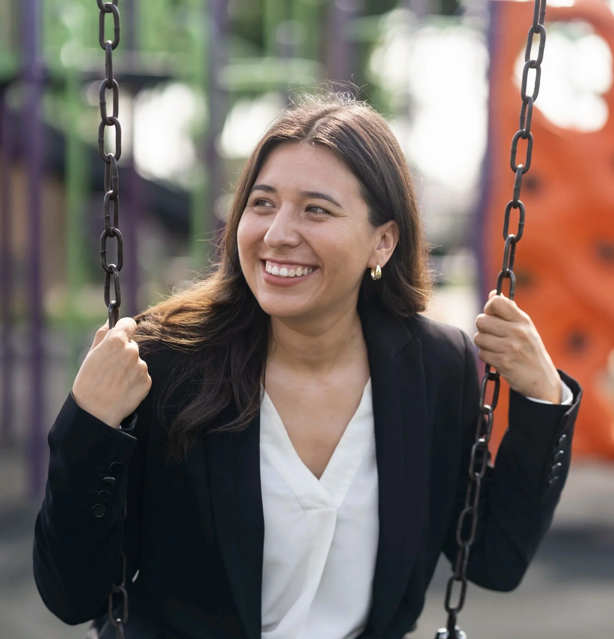 A woman smiling and sitting on a swing at a playground, holding onto the swing's chains.