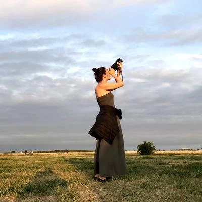 Eine Frau steht in einem offenen Feld, fotografiert die Szenerie mit einer Kamera, Himmel mit Wolken im Hintergrund.